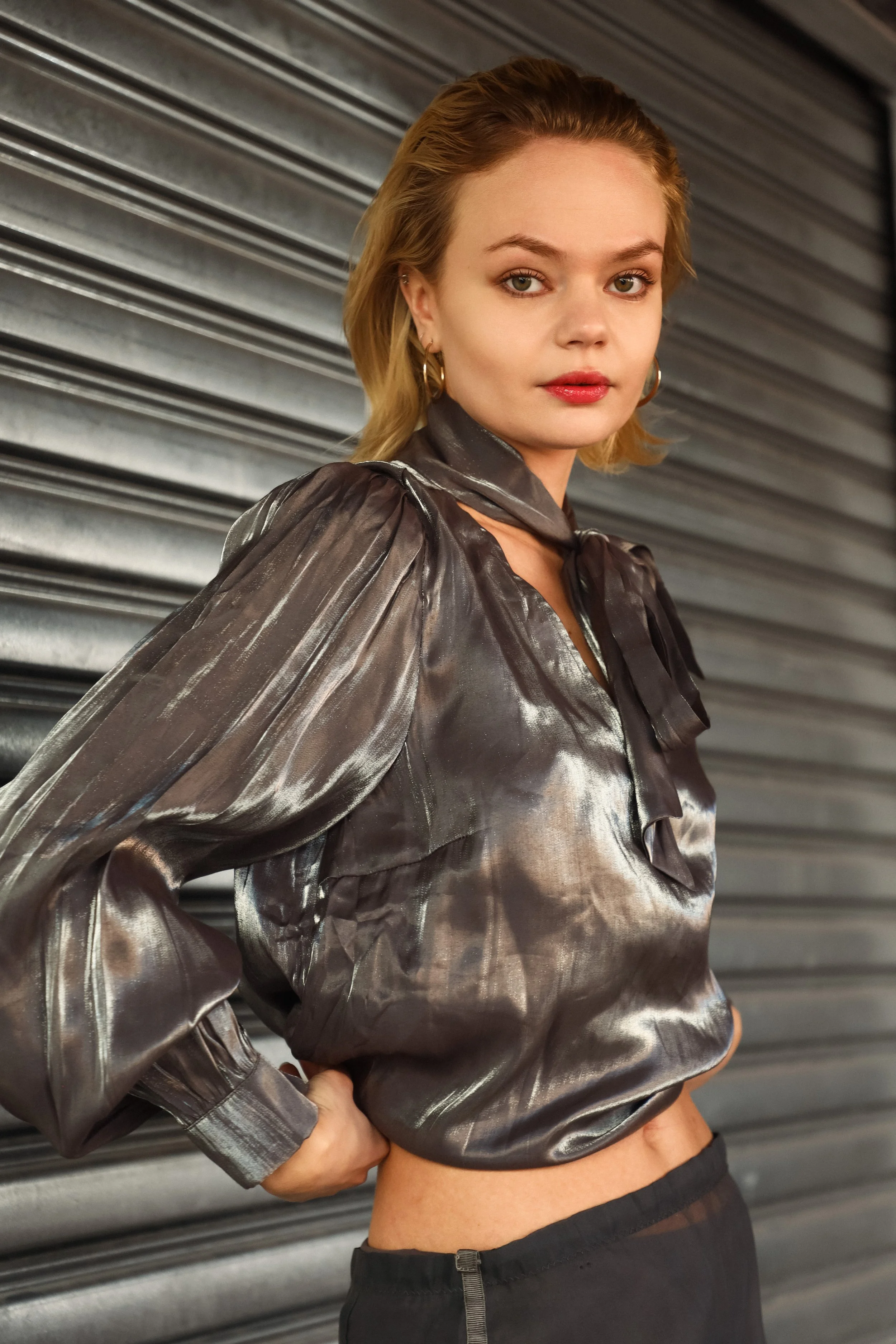 A young woman with blonde hair styled in waves, wearing a metallic dark gray blouse with puffed sleeves and a bow at the collar, standing against a metal shutter background.