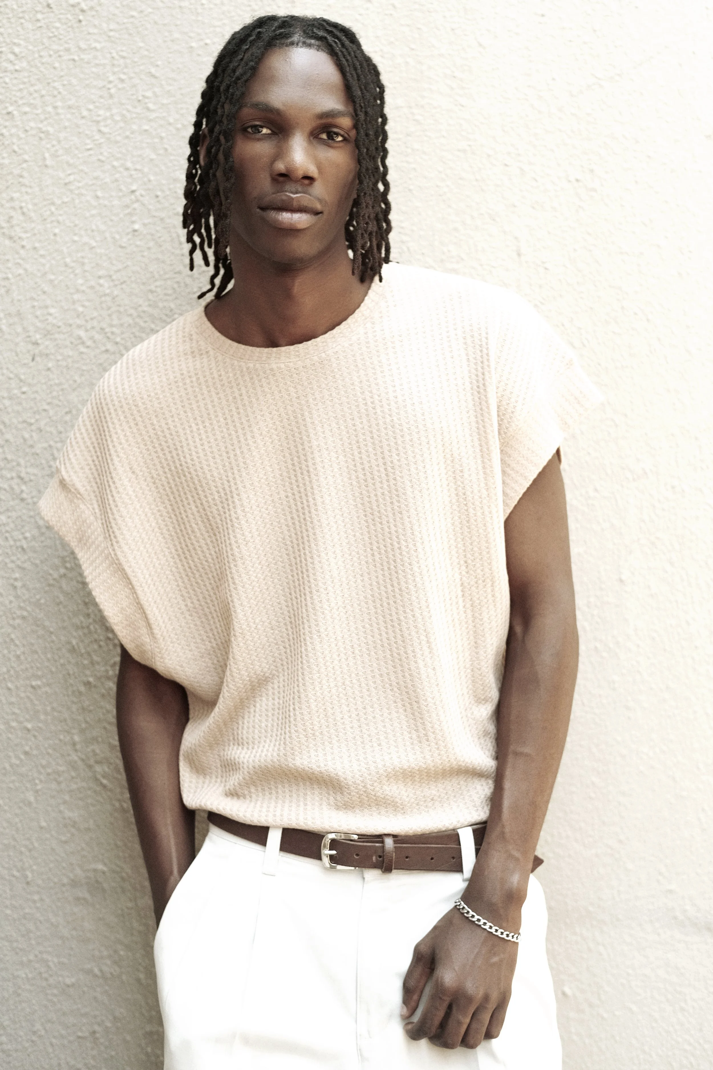 TSHEDZA Ramavheya from AURA Models with dark skin and dreadlocks posing against a textured beige wall, wearing a beige textured t-shirt, white pants, a brown belt, and a silver chain bracelet.