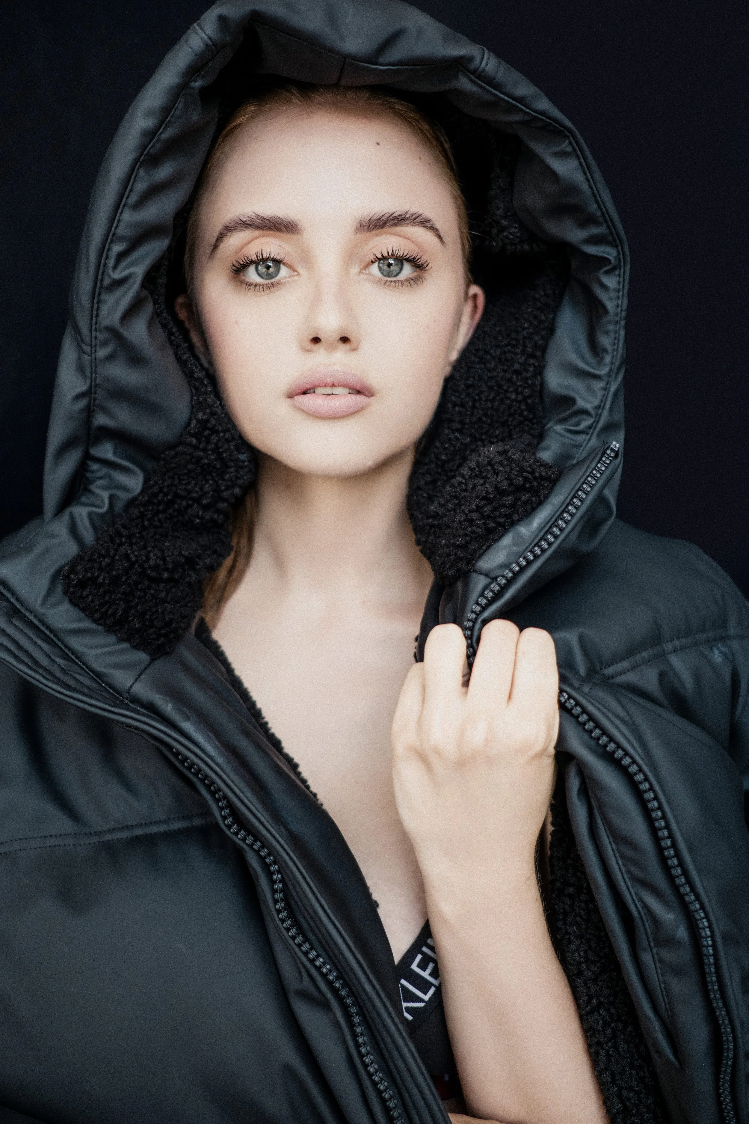 DANIELLE Laubscher from AURA Models with blue eyes and fair skin wearing a black hooded jacket with a fur-lined collar, holding the zipper near her face, against a dark background.