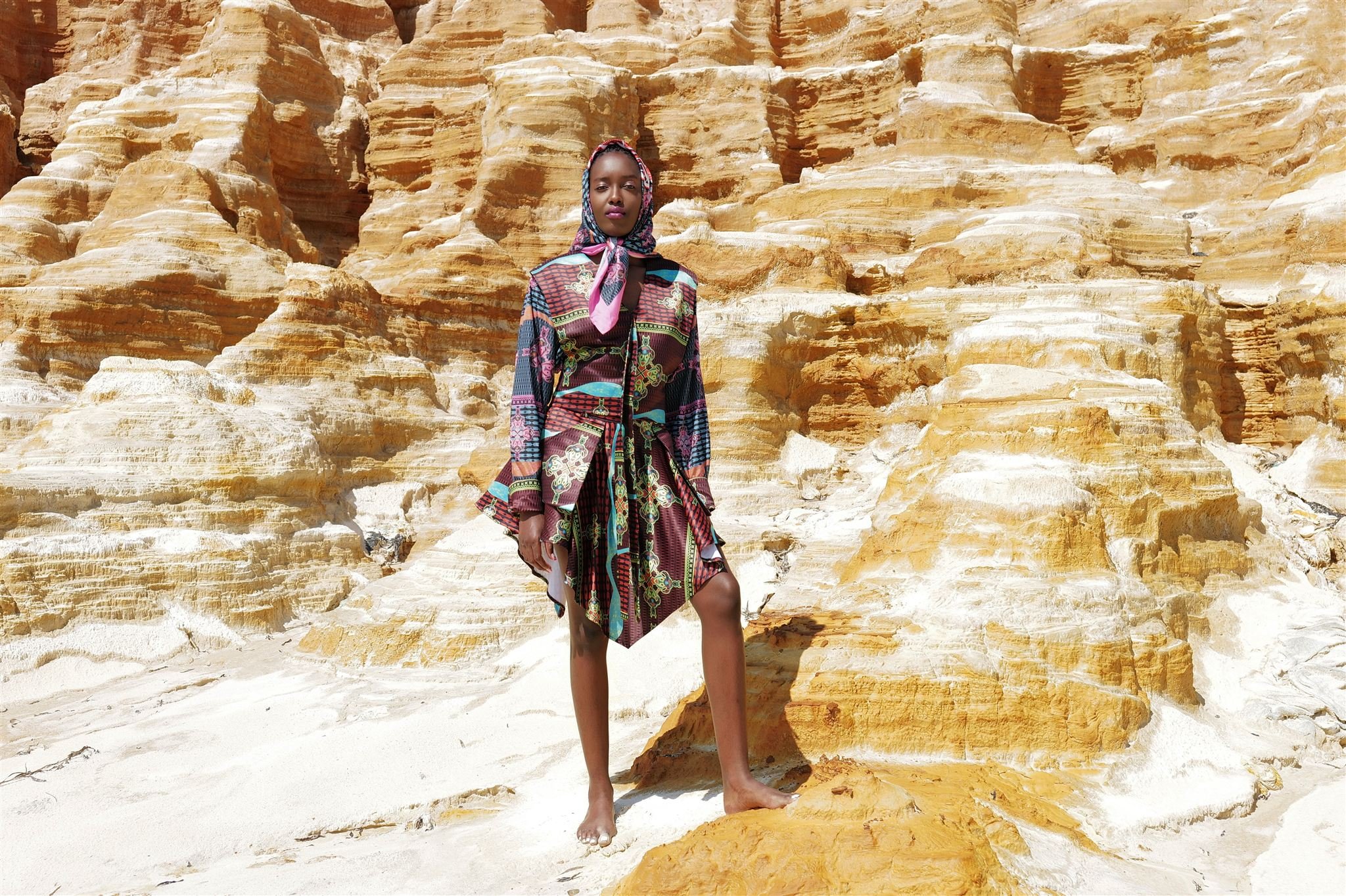 KIM Chakanyuka from AURA Models standing barefoot on a sandy area in front of a large, layered sandstone formation, wearing a colorful, patterned Masamara dress and a matching headscarf.