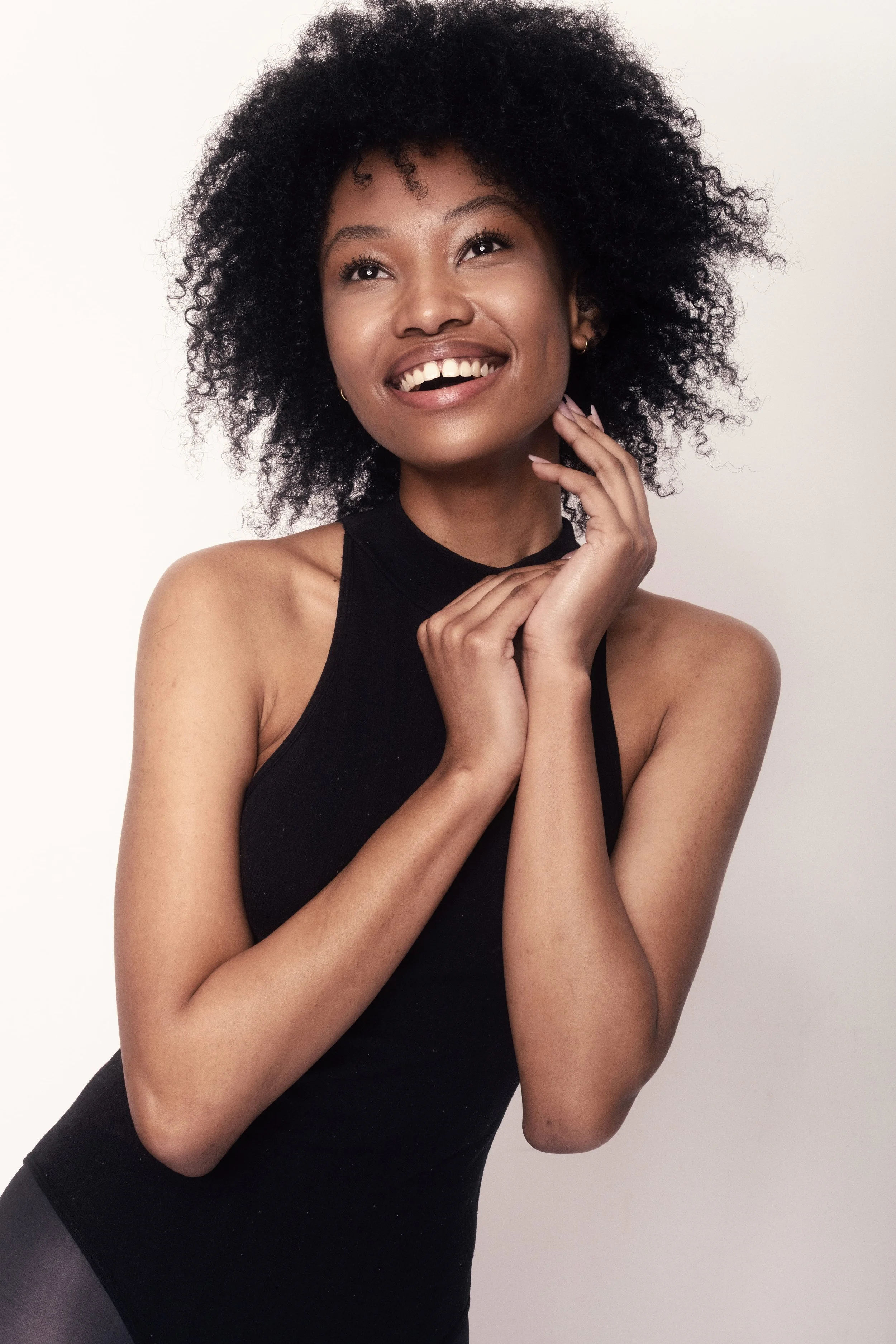 QUISHA Rapoo from AURA Models with curly black hair wearing a black sleeveless top smiling and touching her face with one hand.