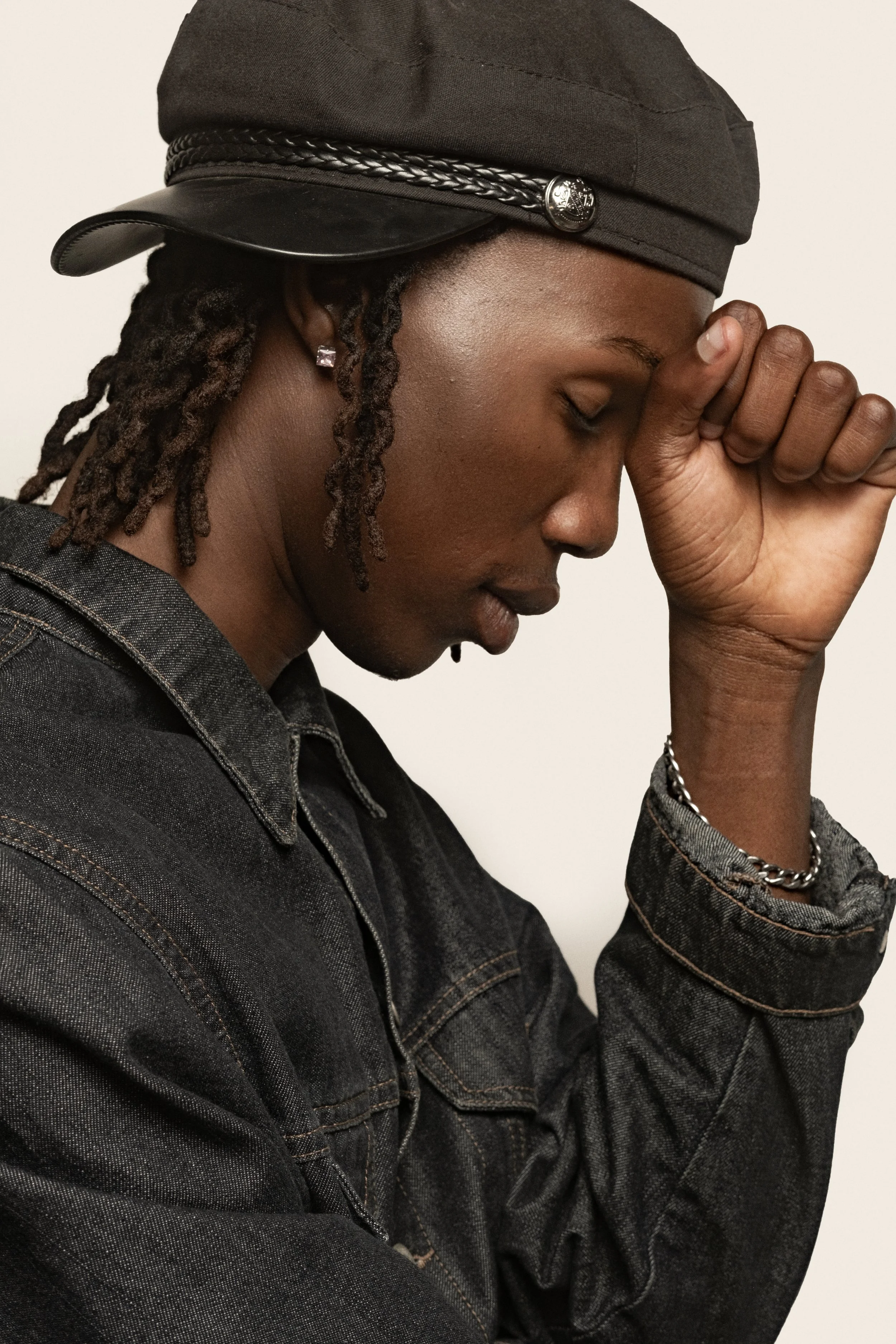 TSHEDZA Ramavheya from AURA Models with dark skin, wearing a black hat and a black denim jacket, has her eyes closed and head bowed, with her right hand touching her forehead.