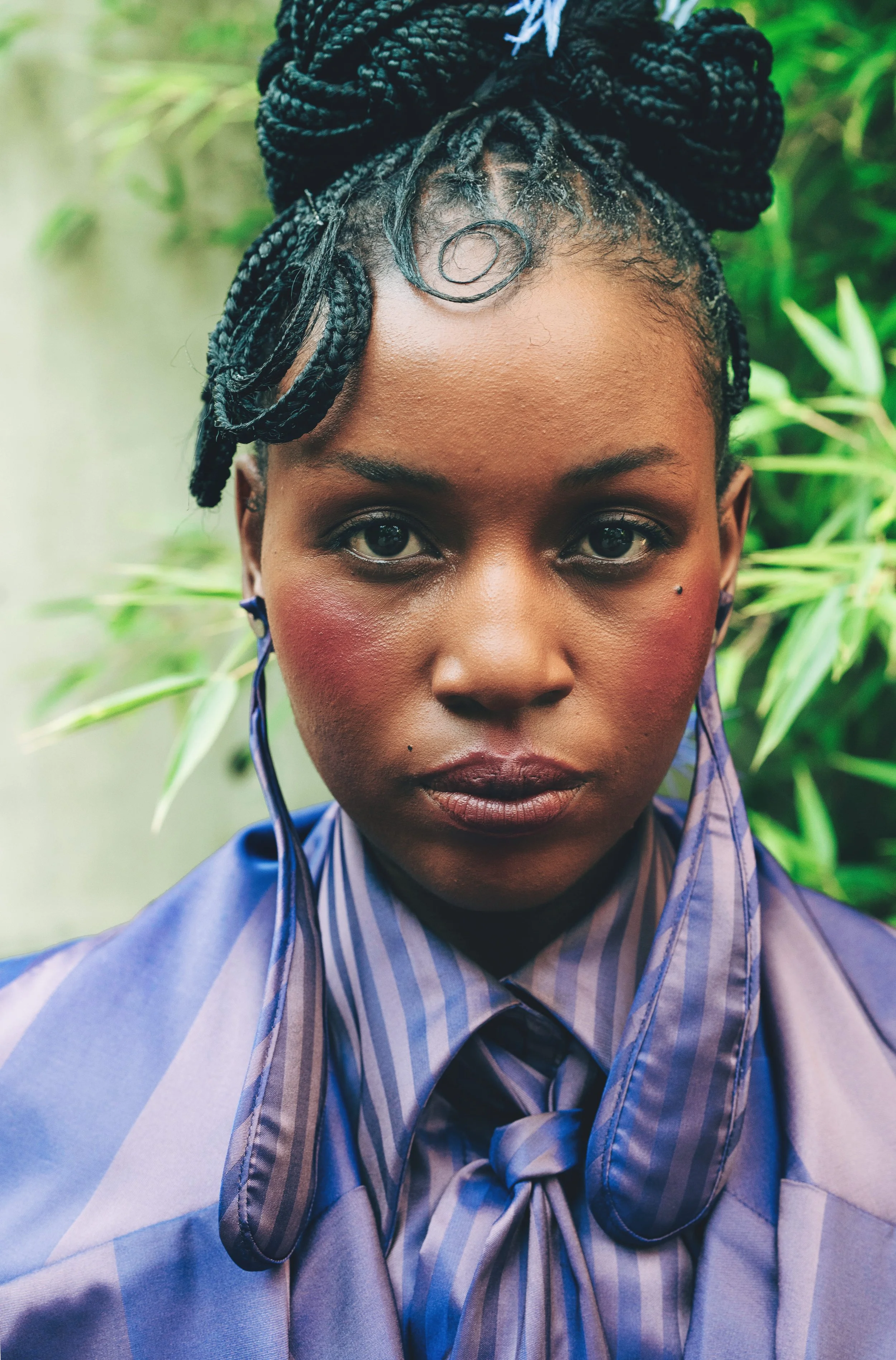 Close-up of RIVO Baloyi from AURA Models with braided hair styled in an updo, wearing a striped shirt and a matching tie, with a blurred green background.