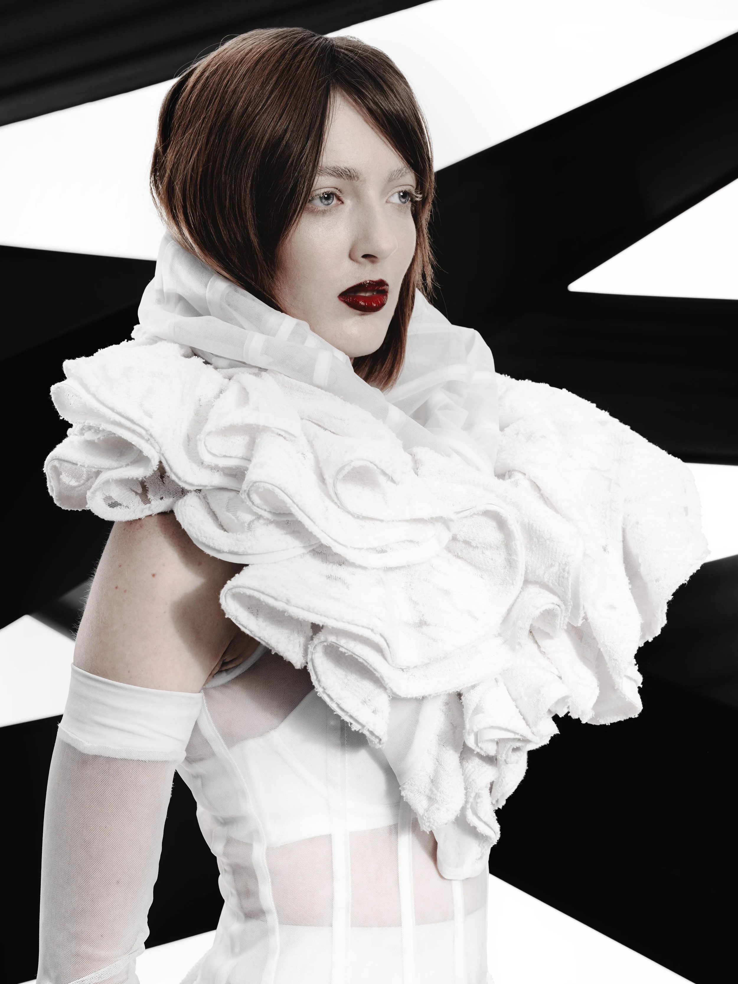 CAYLEIGH Bancroft from AURA Models with short, bobbed brown hair and pale skin wearing a white dress and sheer gloves, holding a large white ruffled fabric. The background features black and white geometric shapes.