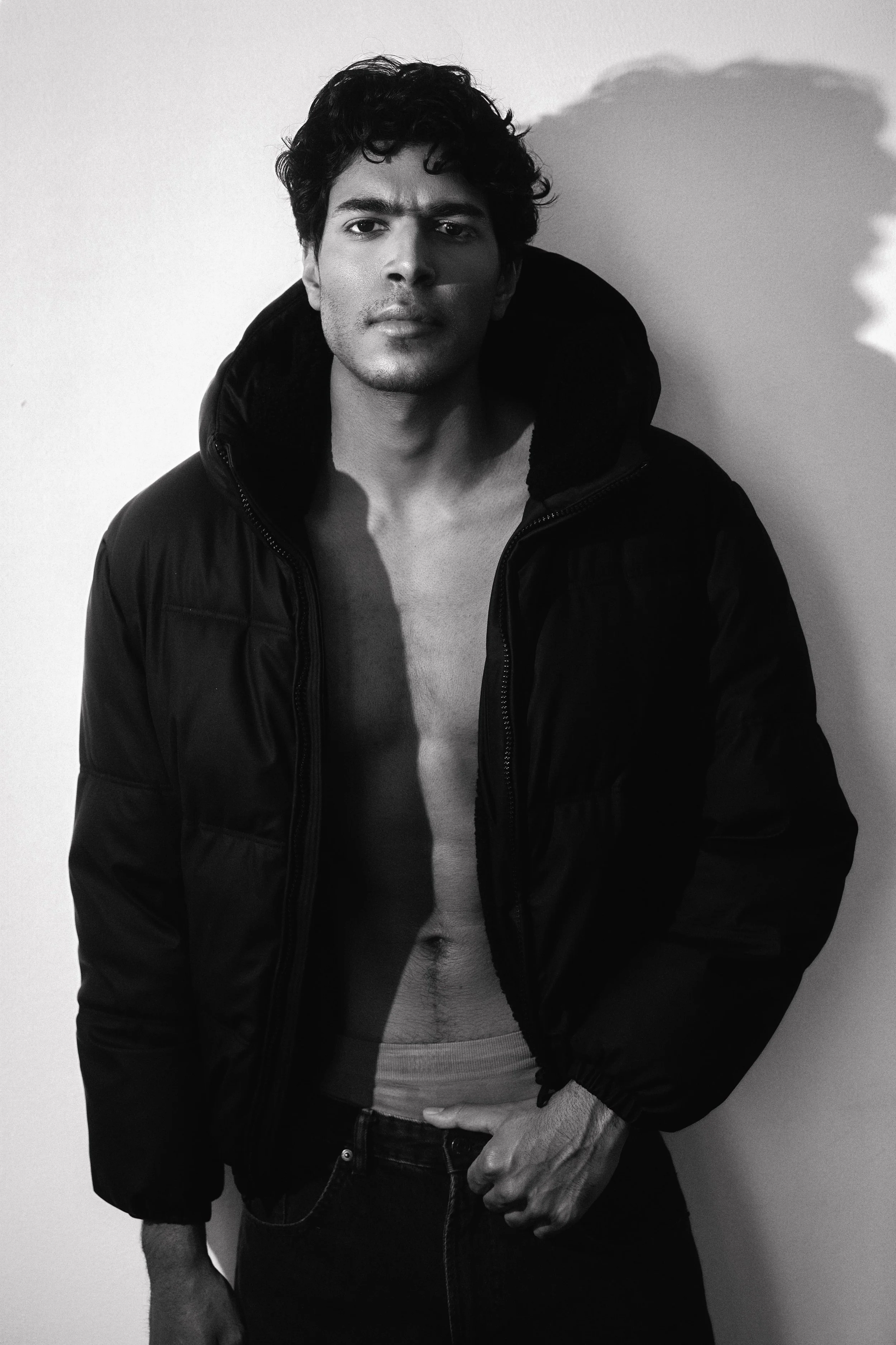Black and white photo of CALEB Naidoo from AURA Models wearing a black jacket with a hood, shirtless underneath, standing against a plain wall. His right hand is in his pocket, and he is looking directly at the camera.