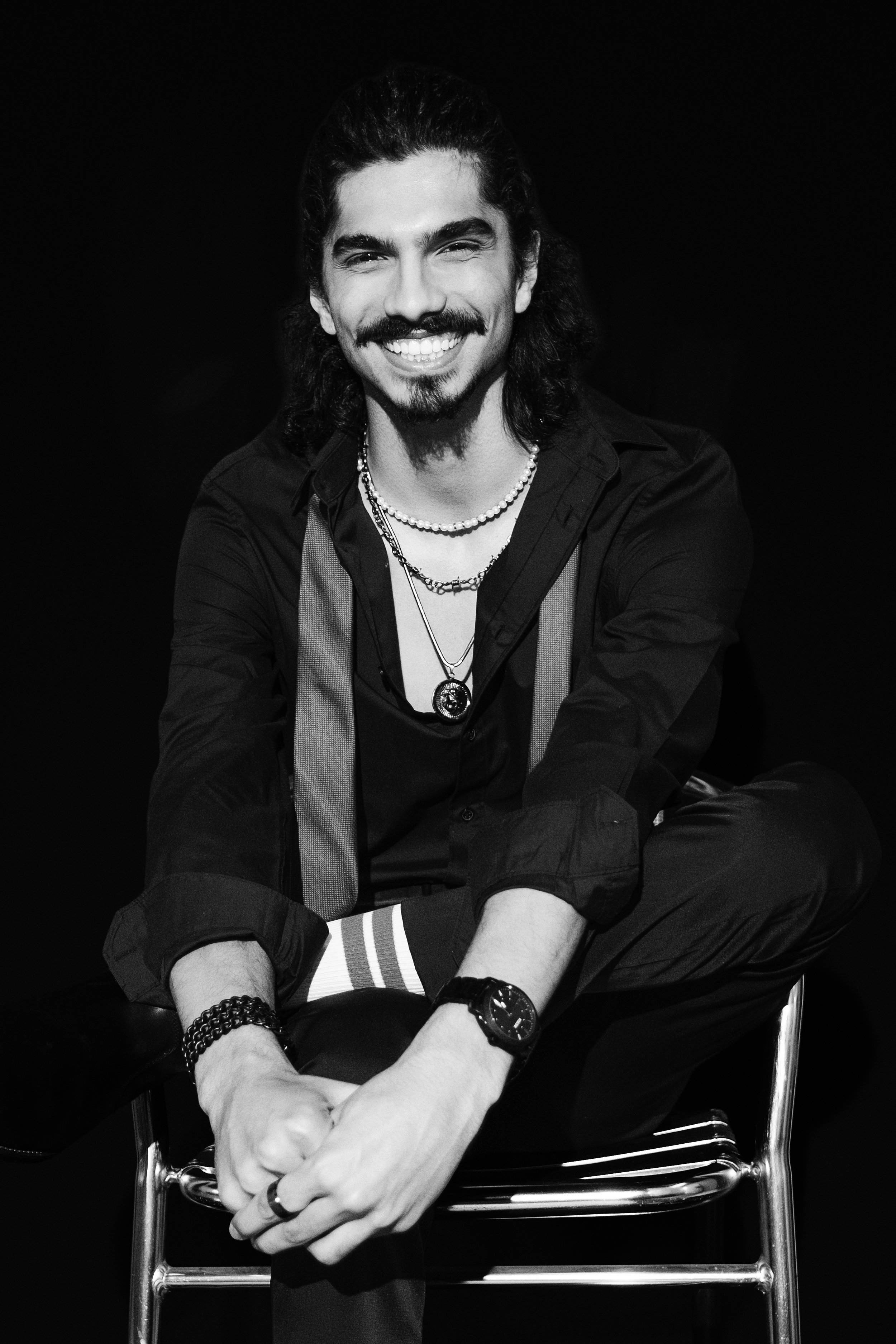 Black and white photo of a smiling man with long dark hair, beard, mustache, wearing layered necklaces, a watch, and casual clothing, sitting cross-legged on a stool against a dark background.