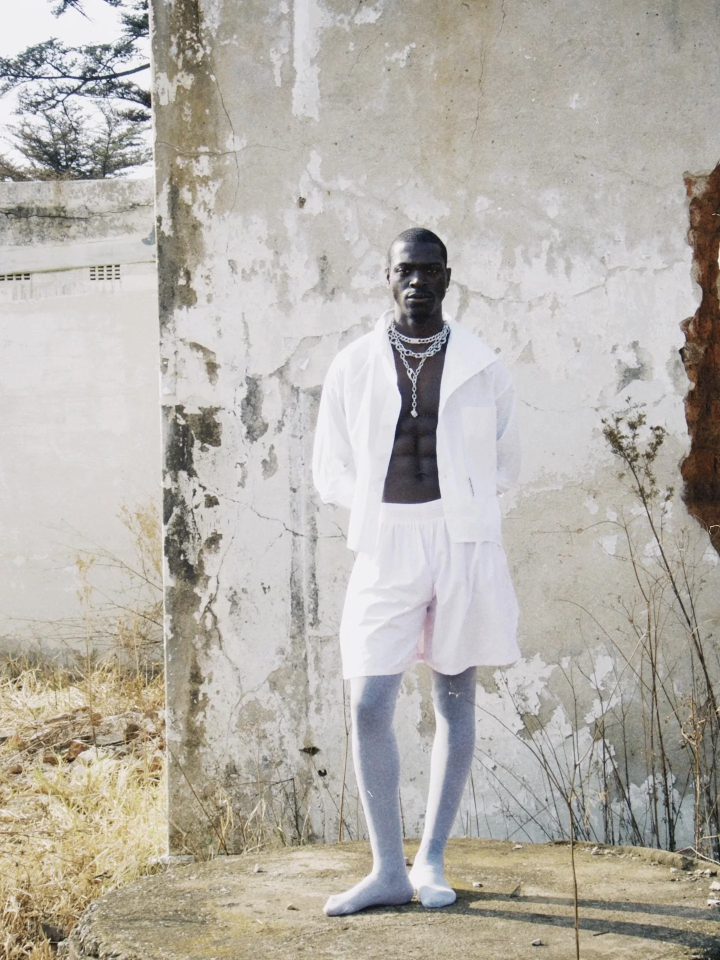 A man dressed in white, wearing chain necklaces, standing outdoors against a weathered wall with some dried grass around.