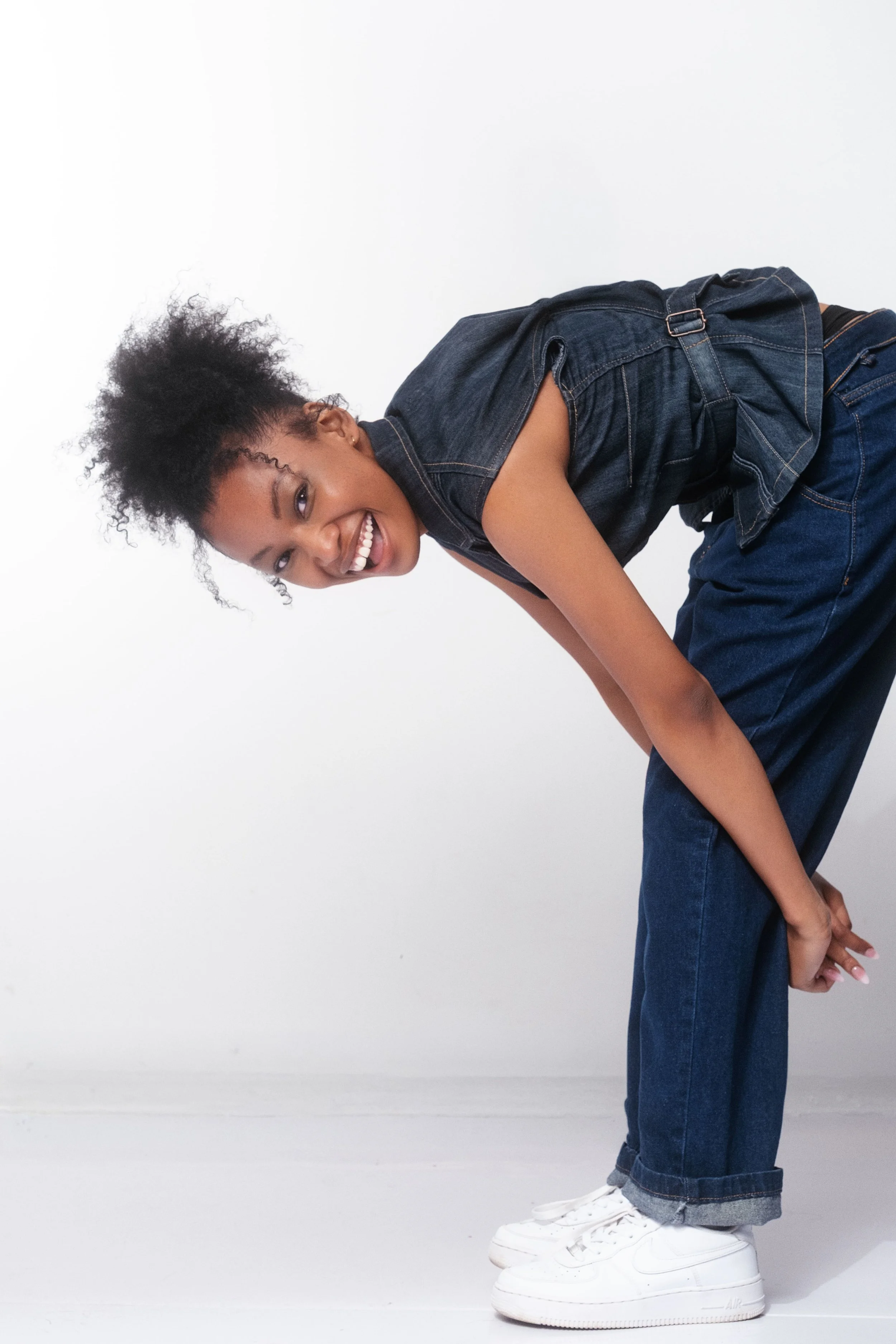 QUISHA Rapoo from AURA Models wearing a sleeveless denim vest and wide-legged jeans, stands against a plain white background. She is smiling and leaning forward with her hands clasped behind her back, wearing white sneakers.