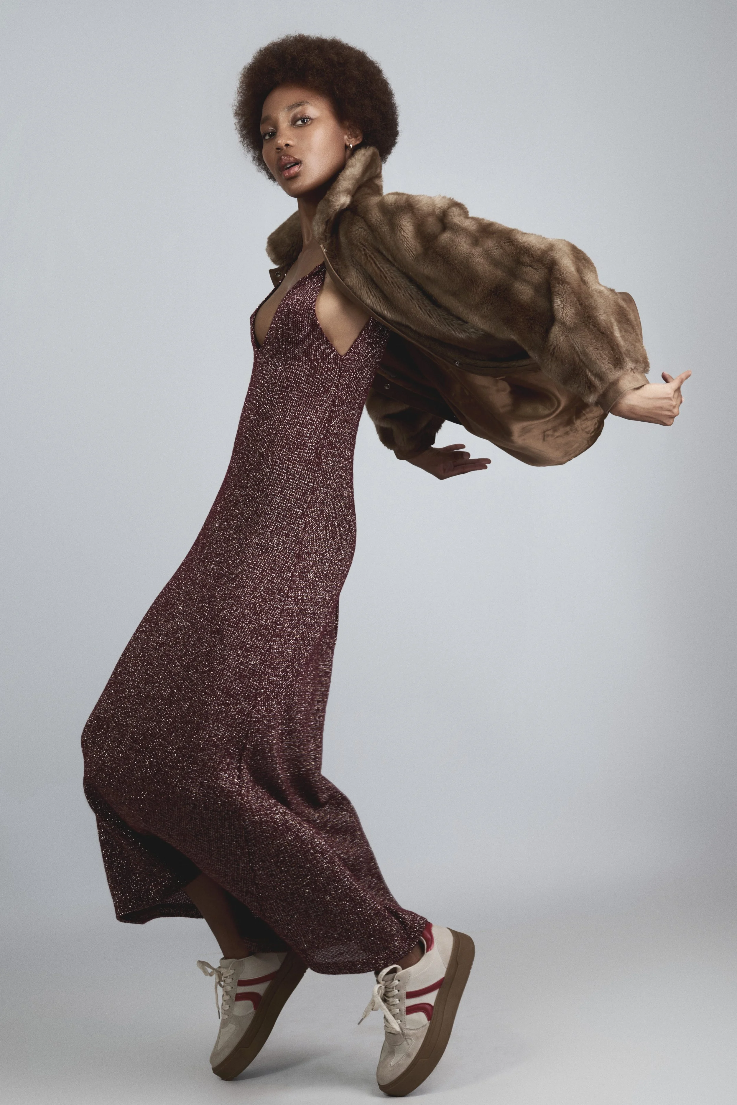 RORY Merafe from AURA Models with curly hair wearing a shimmering red dress, a brown fur coat, and sneakers poses against a plain background.