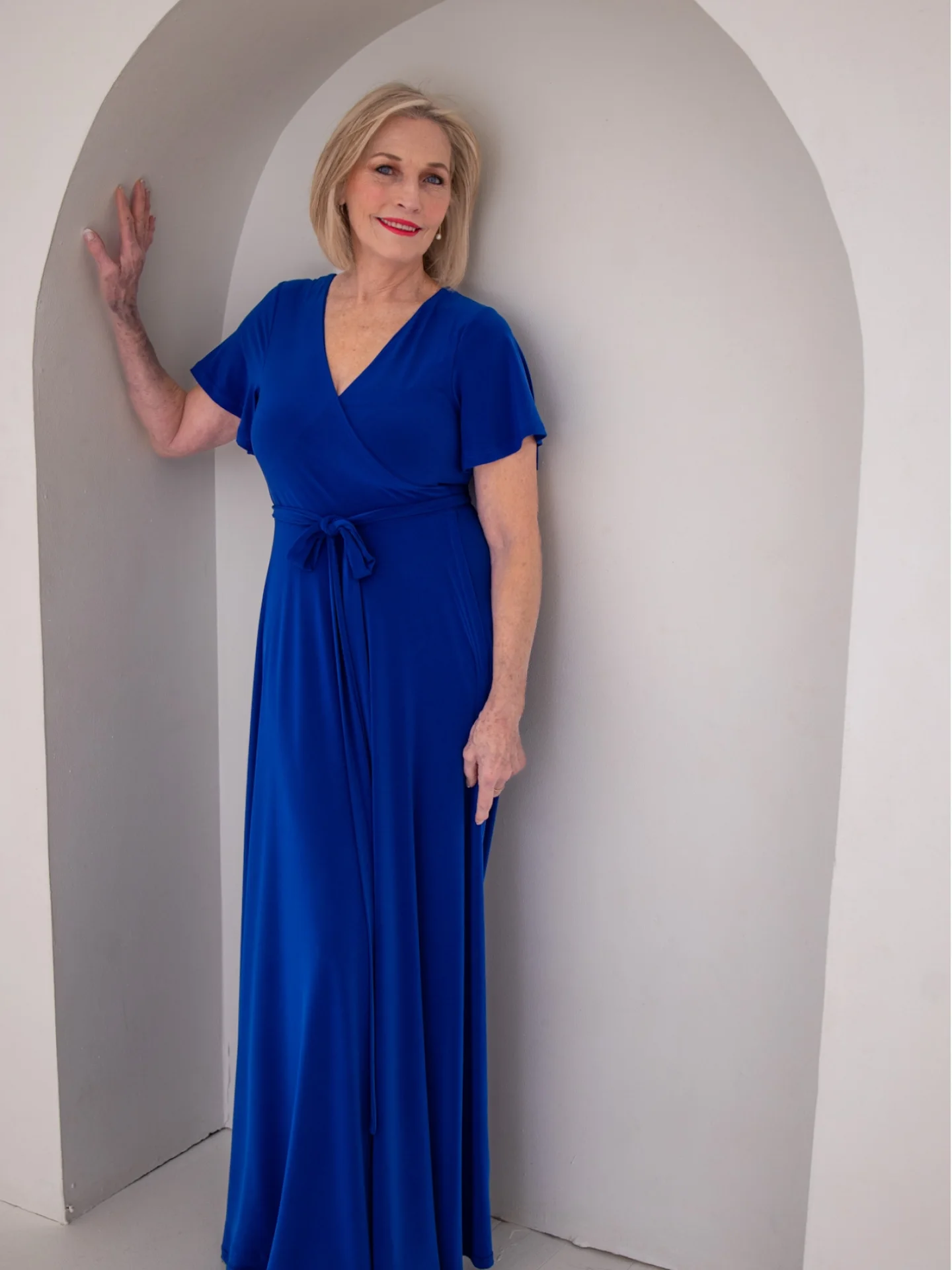 MARIETA Ferreira from AURA Models in a vibrant blue dress standing comfortably in an arched niche in a white wall, smiling at the camera.