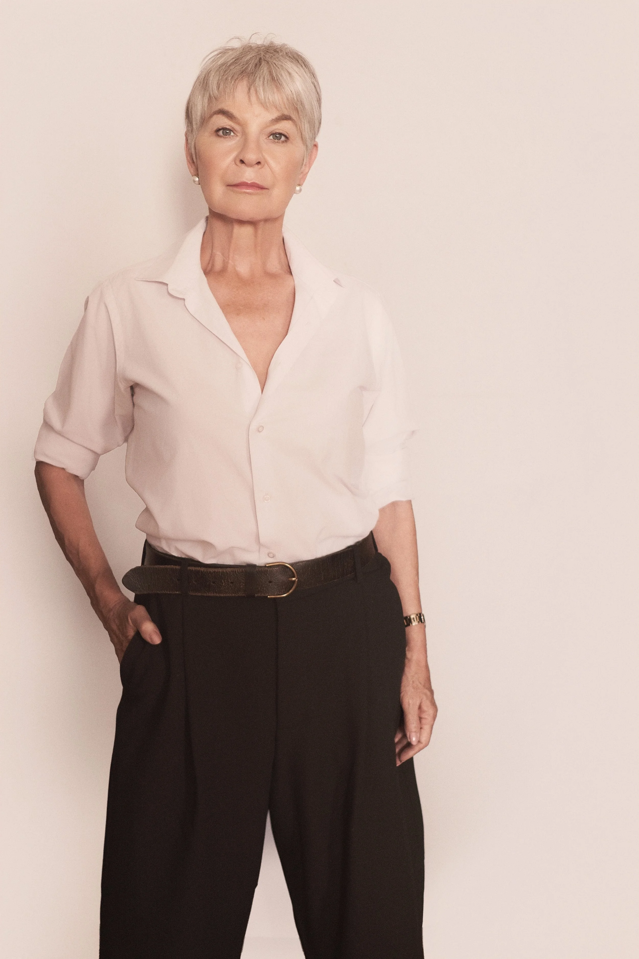 BRIDGET van Zyl from AURA Models with short gray hair, wearing a white button-up shirt with the sleeves rolled up and black pants with a belt, standing against a plain white wall.