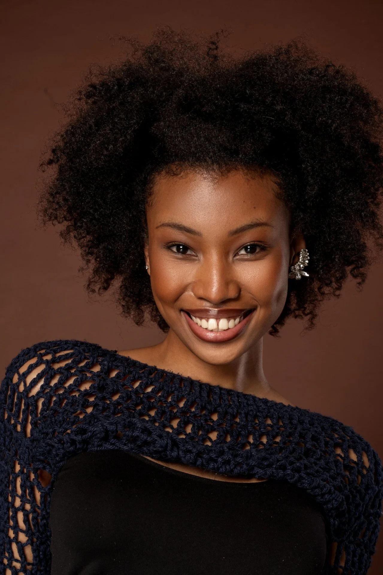 Portrait of a young African American woman with curly hair, smiling, wearing a navy crocheted top and statement earrings against a brown background.