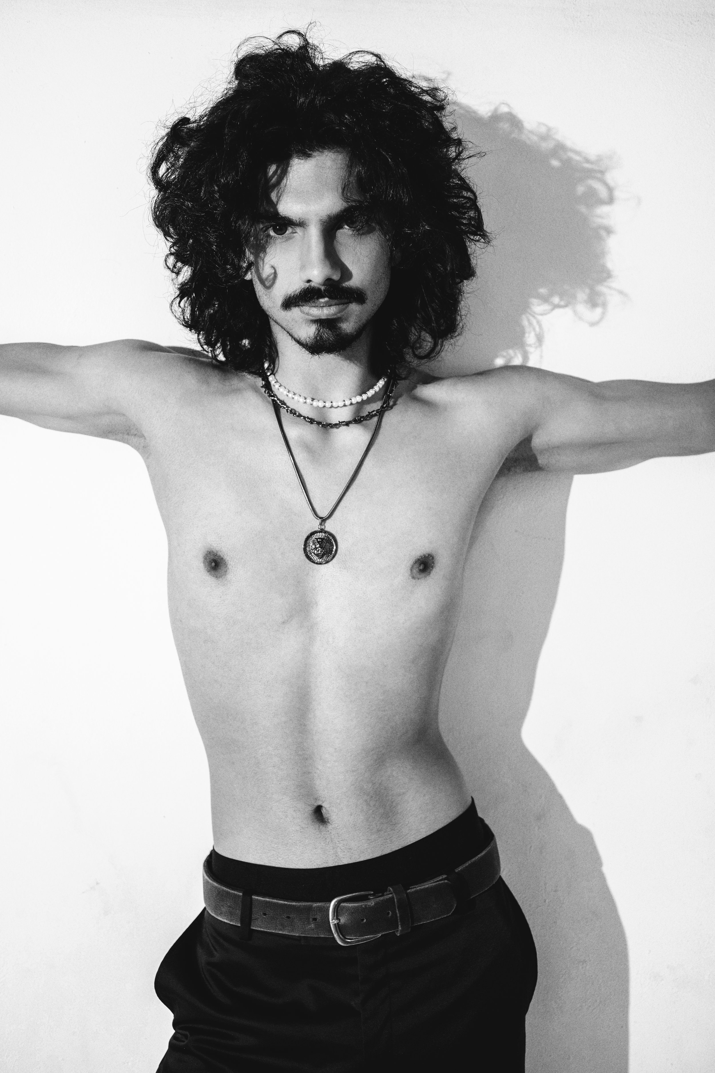 A black-and-white photo of a shirtless man with wild curly hair, wearing multiple necklaces and black pants with a belt, standing against a white background.
