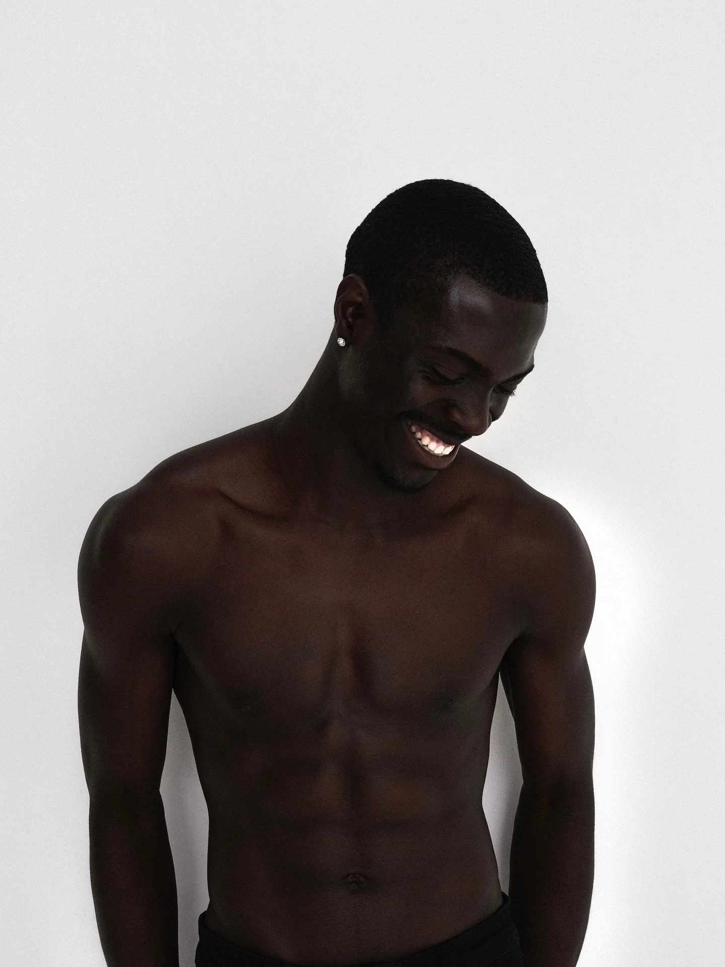 A young Black man with short hair and a bright smile looking down against a plain white background.