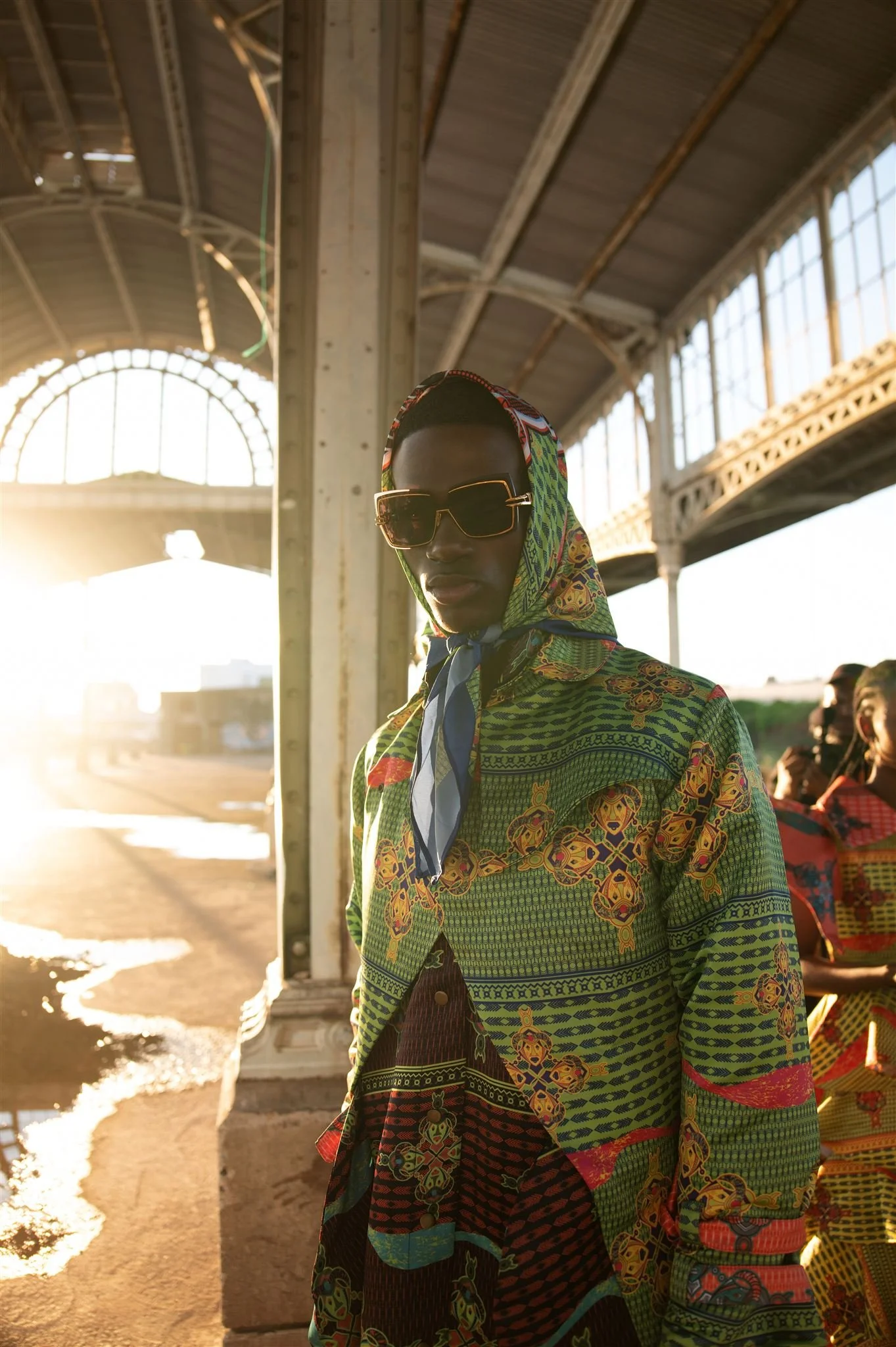 MBHURI Baloyi from AURA Models wearing oversized sunglasses and a Masamara hooded, colorful, patterned outfit standing under a large industrial-looking structure with sunlight in the background.