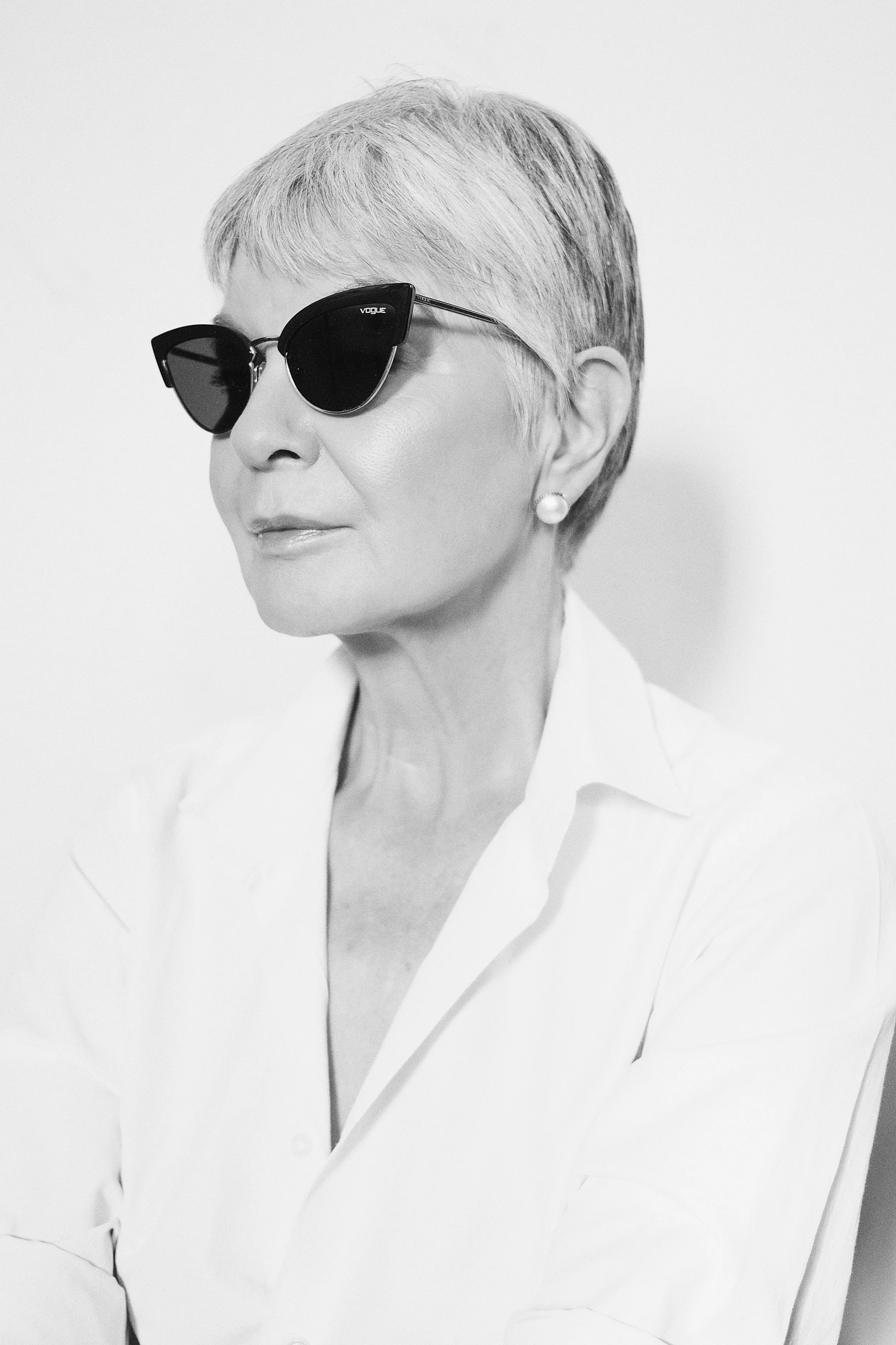 A black-and-white photo of BRIDGET van Zyl from AURA Models with short hair, wearing dark sunglasses, pearl earrings, and a white shirt with a collar, looking to her left with a calm expression.