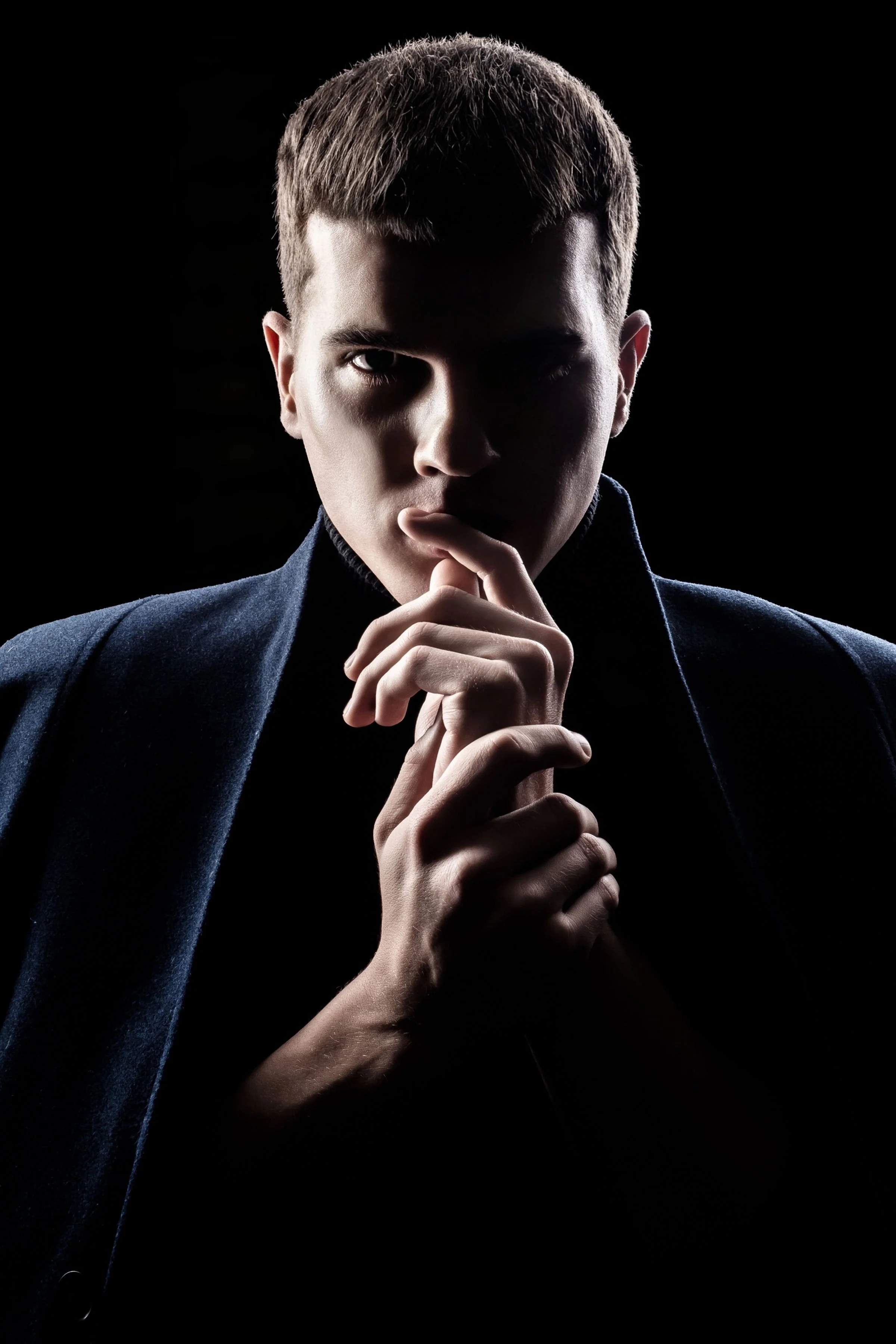 A young man with short hair and light skin holding his fingers to his lips, wearing a dark coat against a black background.