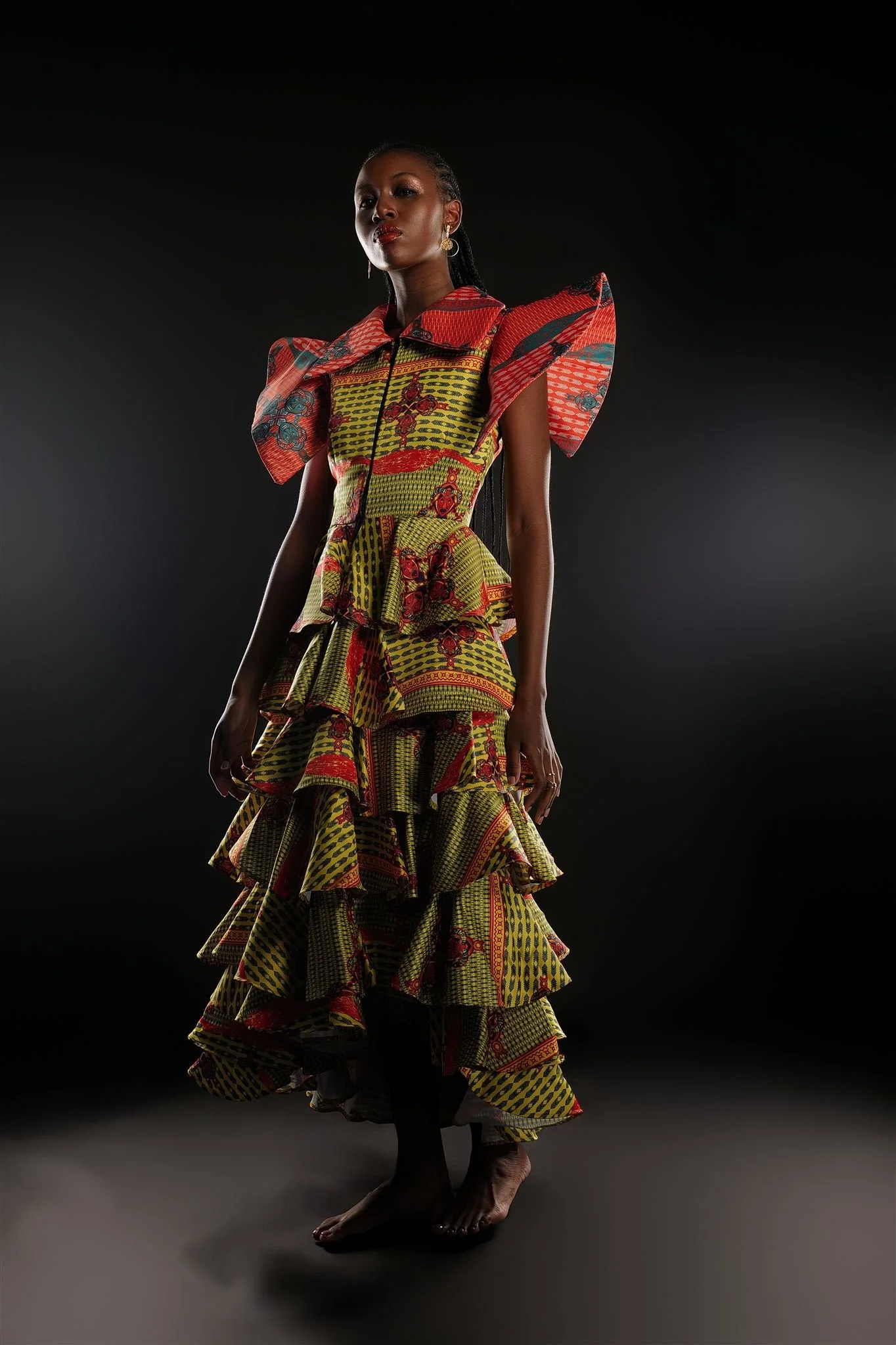 RIVO Baloyi from AURA Models wearing a colorful Masamara African print dress with layered ruffles and puffed sleeves, standing barefoot against a dark background.