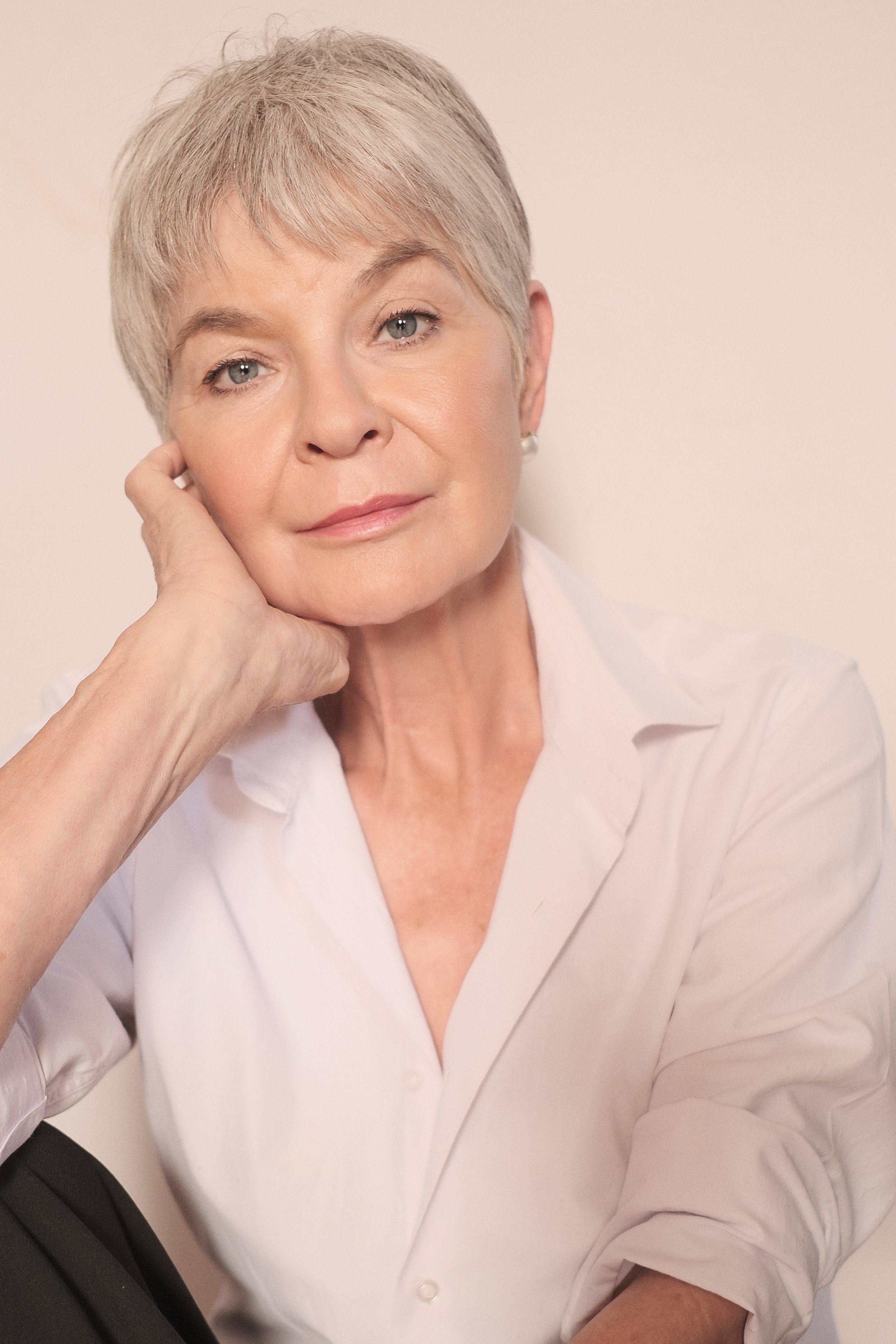 Portrait of BRIDGET van Zyl from AURA Models with short gray hair, wearing a white shirt, resting her chin on her hand, looking thoughtfully at the camera.