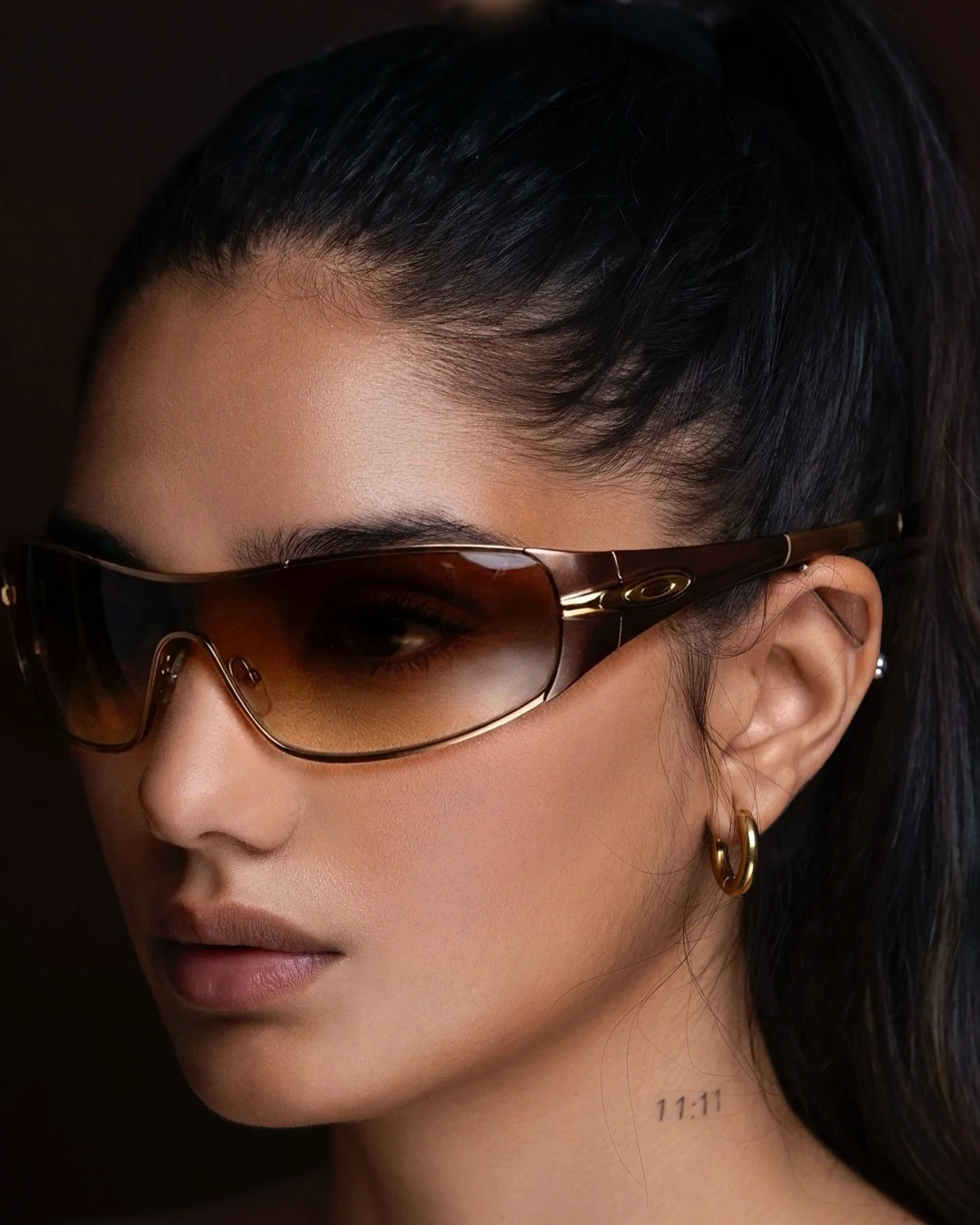 Close-up of EMILY Francis from AURA Models wearing large sunglasses, gold hoop earrings, and a small tattoo of '11:11' on her neck.