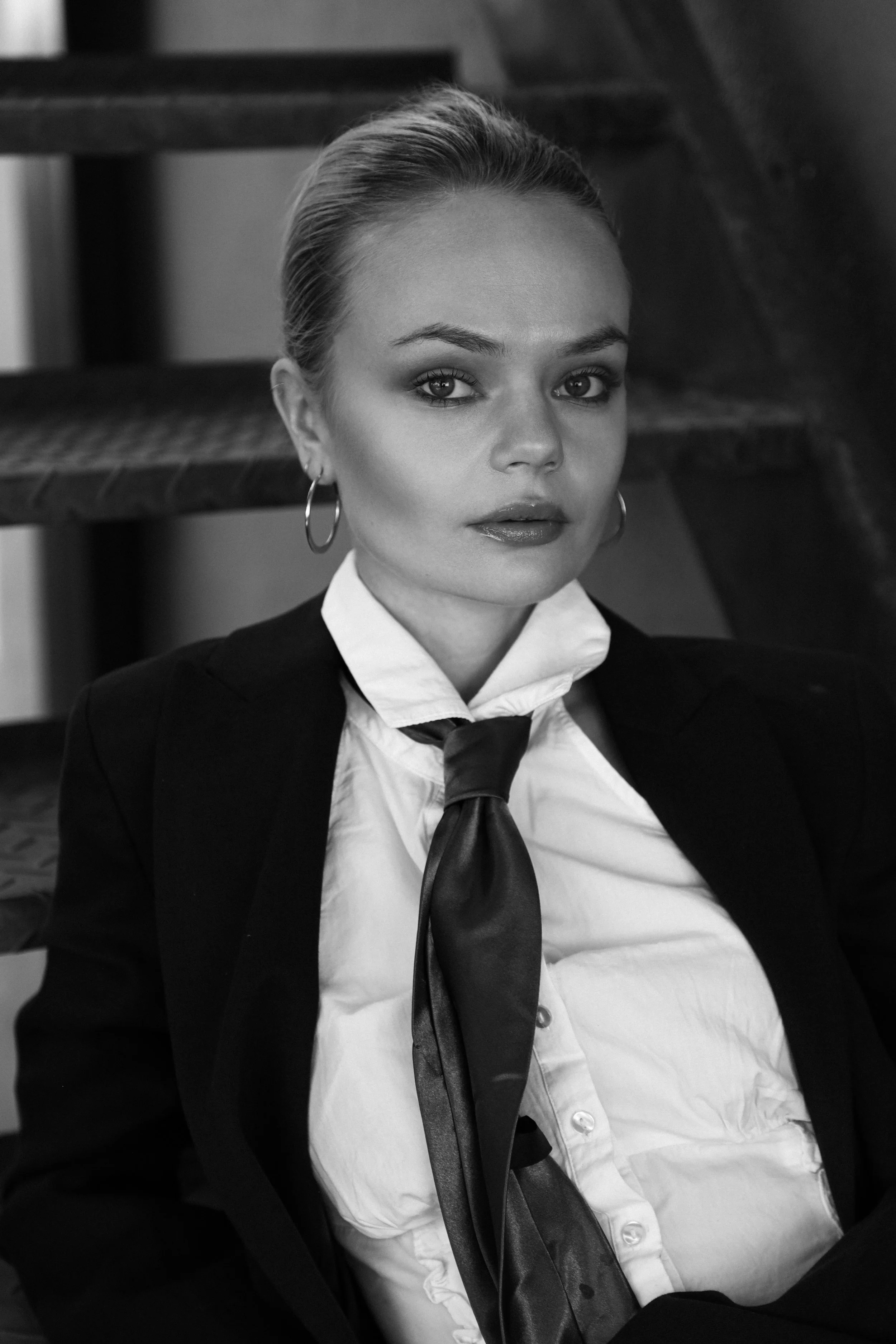 A black-and-white portrait of a woman with short hair, wearing hoop earrings, a white shirt, a black blazer, and a shiny black tie, sitting on stairs.