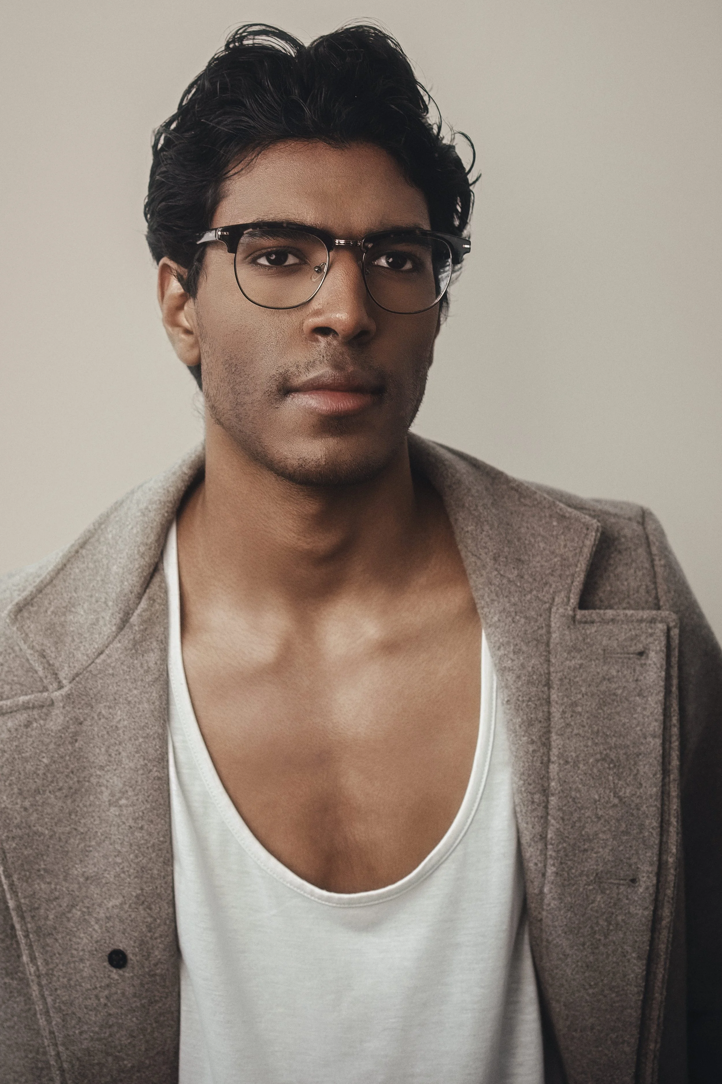 CALEB Naidoo from AURA Models with dark, wavy hair wearing glasses, a white V-neck shirt, and a beige blazer, looking at the camera against a plain background.