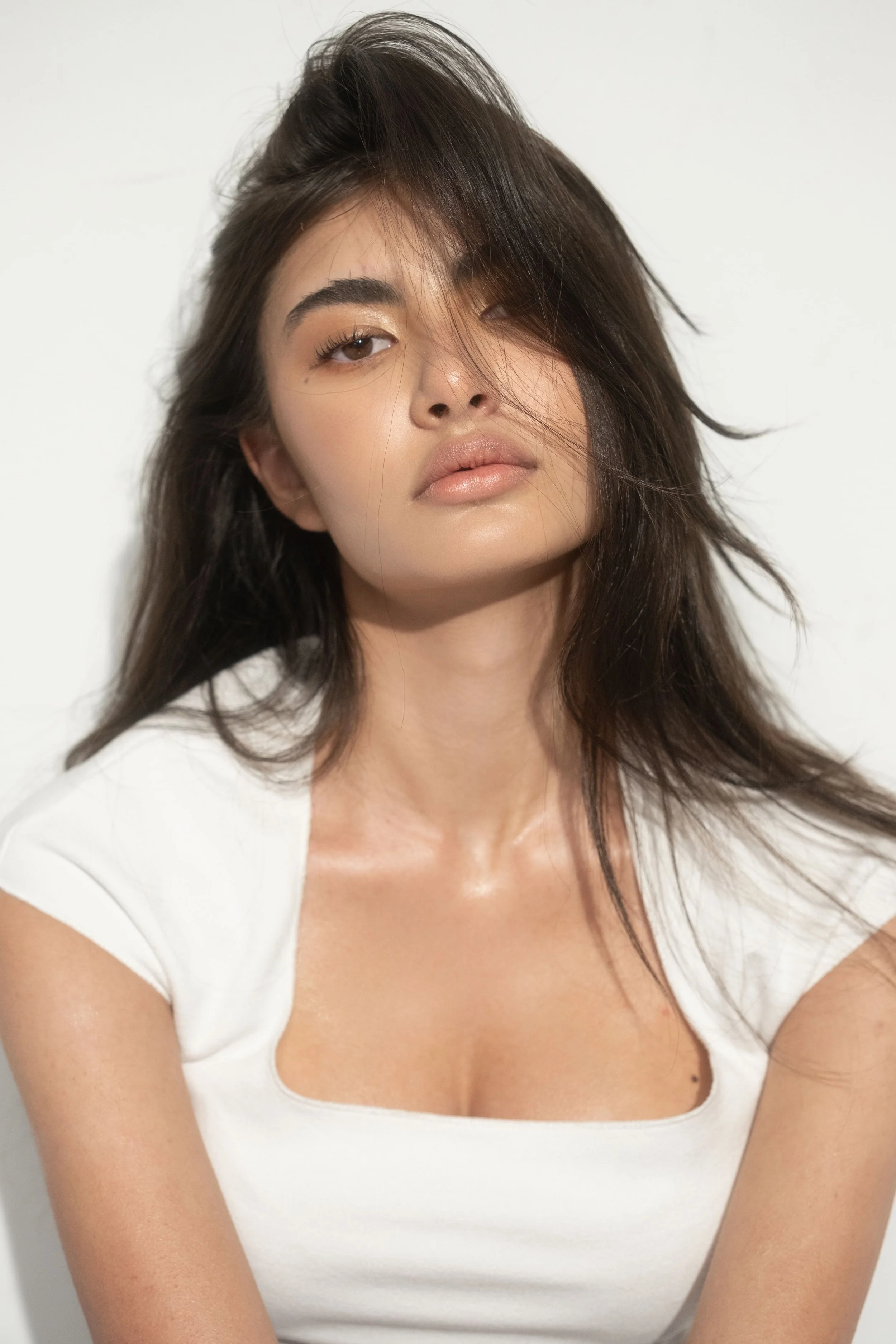 KEIRA Louw from AURA Models with dark, slightly wavy hair partially covering her face, wearing a white shirt with a scooped neckline, standing against a plain light background.