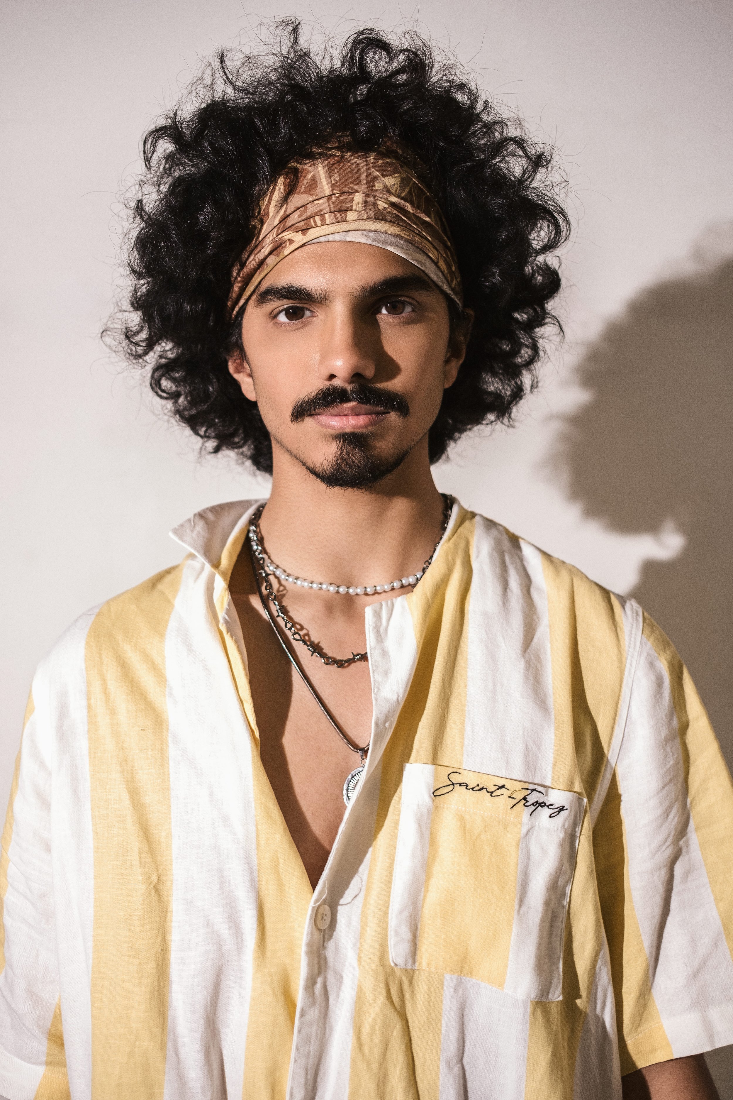 A young man with curly dark hair and a mustache wearing a beige and white striped shirt with a label reading 'Saint Tropez', layered necklaces, and a patterned headscarf, standing against a plain white background with a shadow.