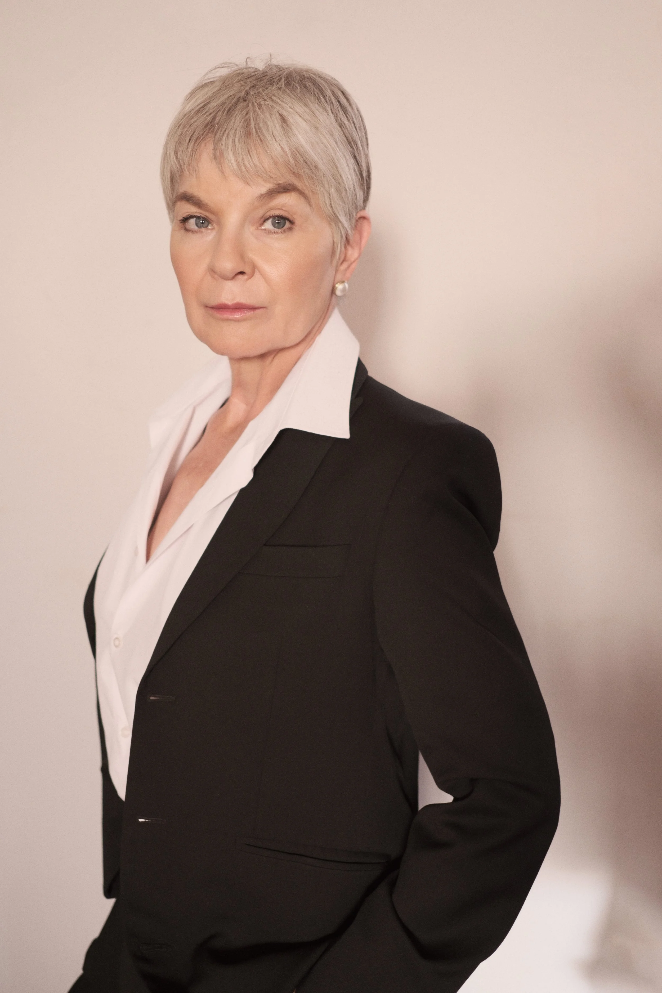 BRIDGET van Zyl from AURA Models with short gray hair, wearing a black blazer over a white shirt, posing against a plain white wall.