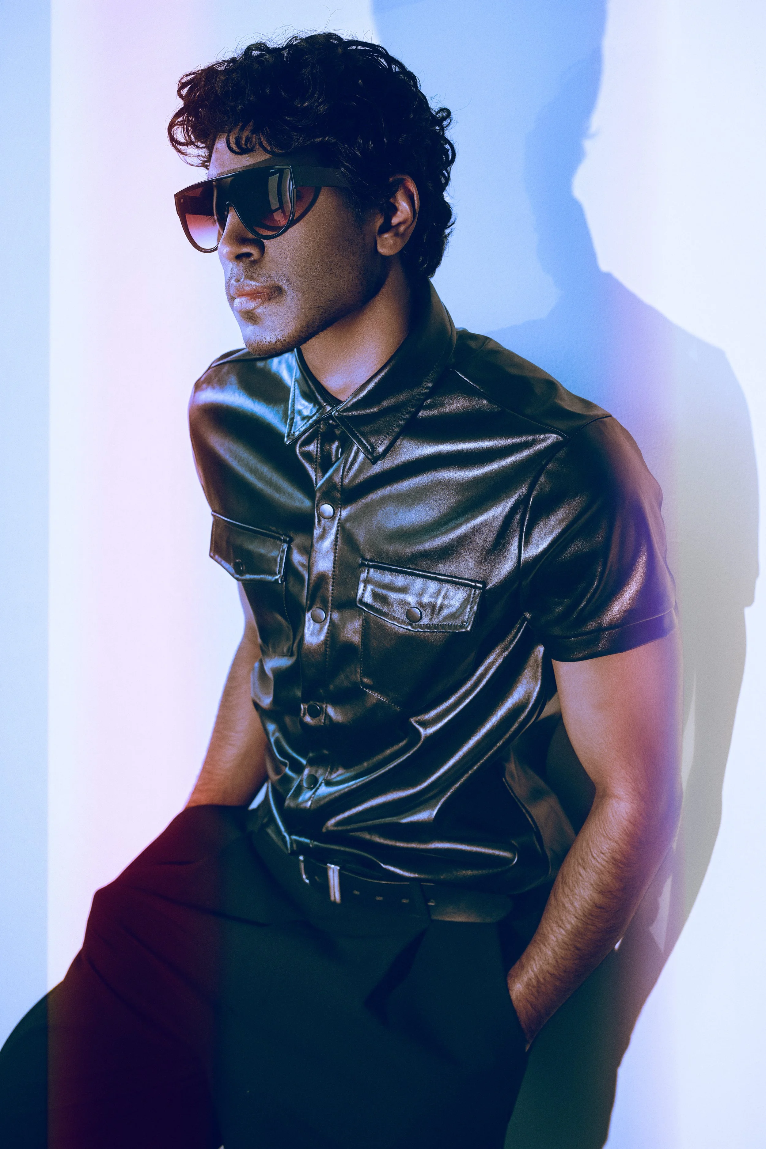 CALEB Naidoo from AURA Models with dark curly hair wearing black sunglasses, a shiny black leather shirt, and black pants, standing against a white wall with shadow.