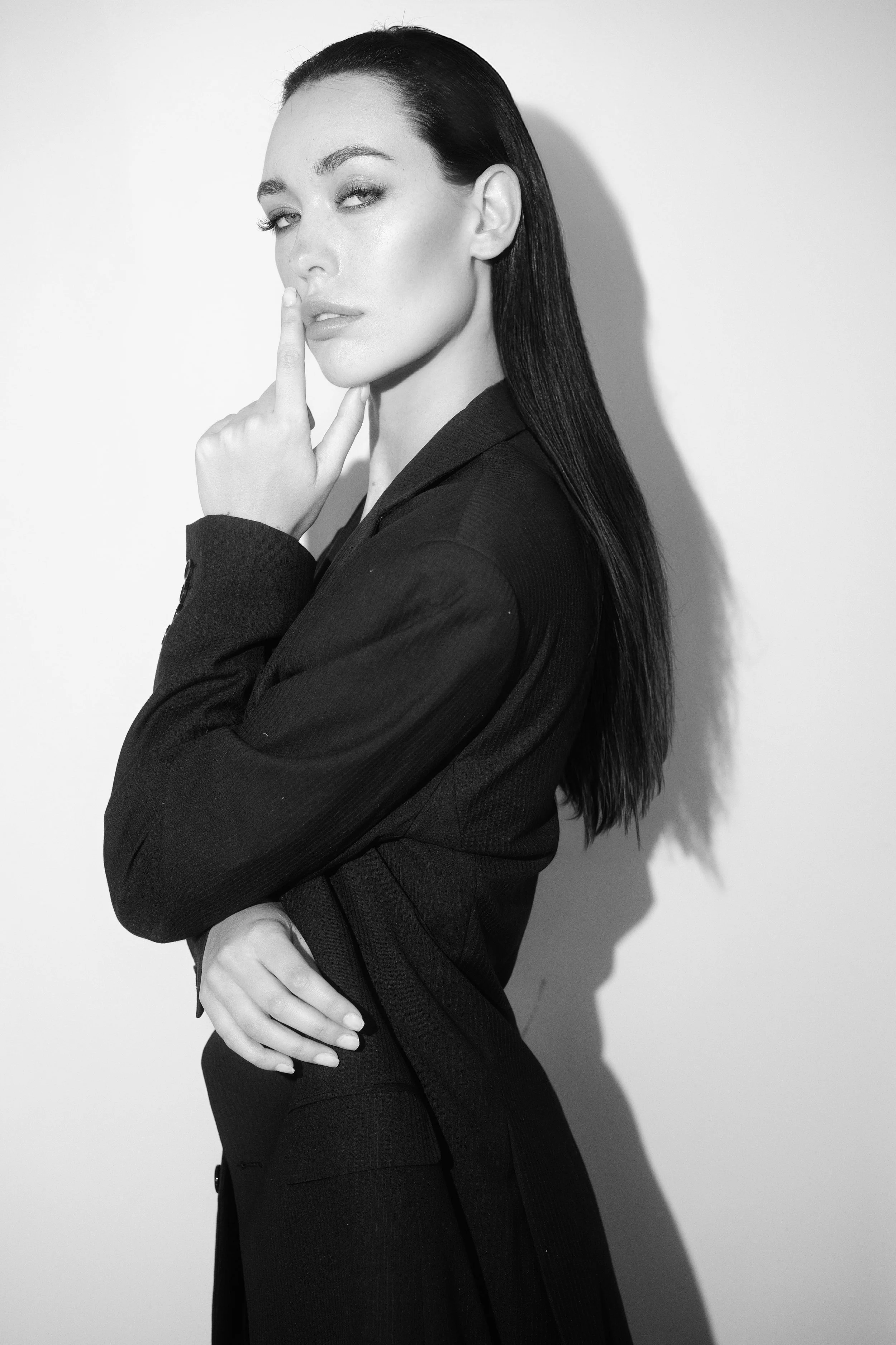Black and white photo of MONIQUE van Loggerenberg from AURA Models with long dark hair, dressed in a suit, making a gesture with her index finger to her lips, standing against a plain background.