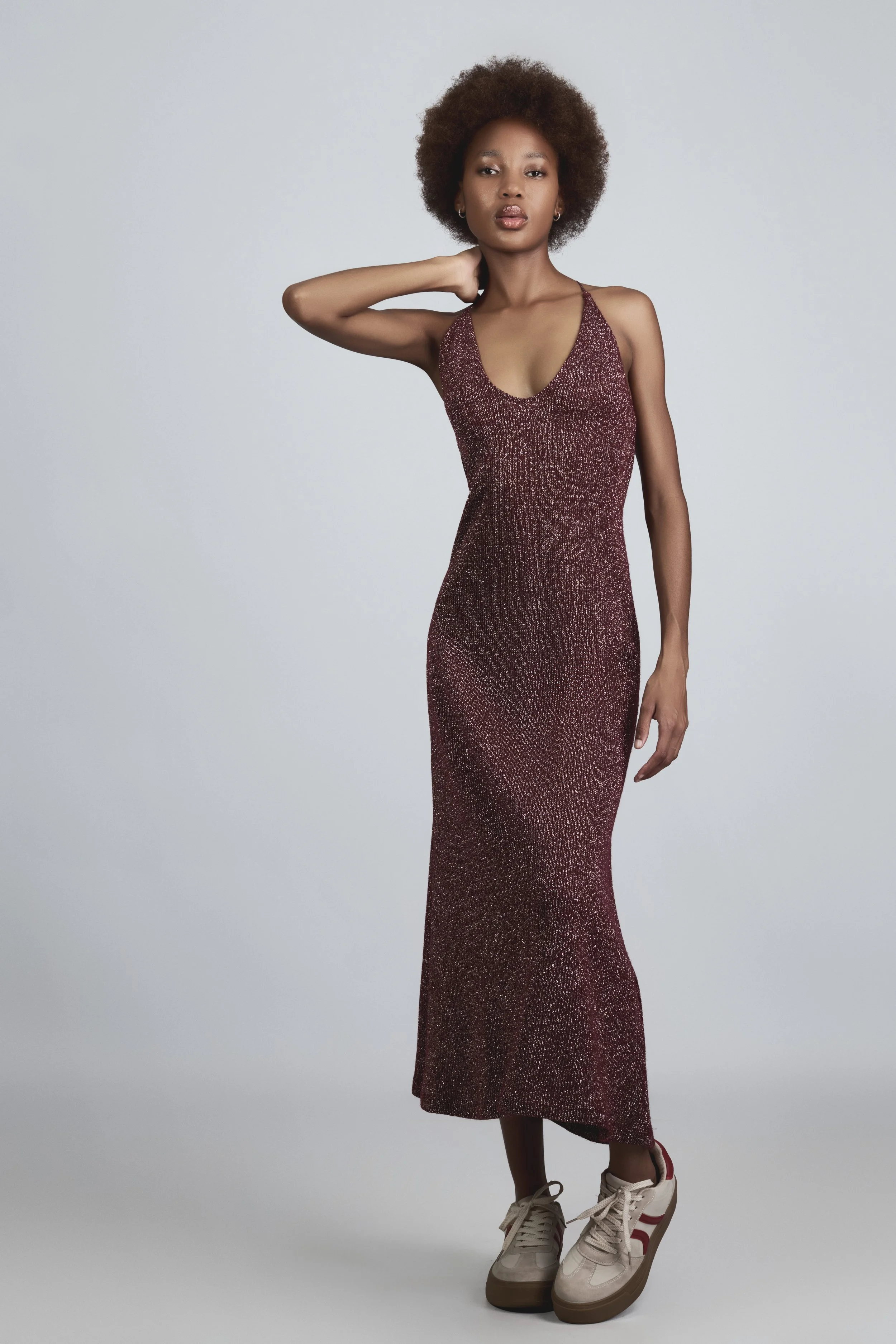 RORY Merafe from AURA Models with an afro hairstyle wearing a long, sparkly, maroon dress and beige sneakers, standing against a plain light gray background.