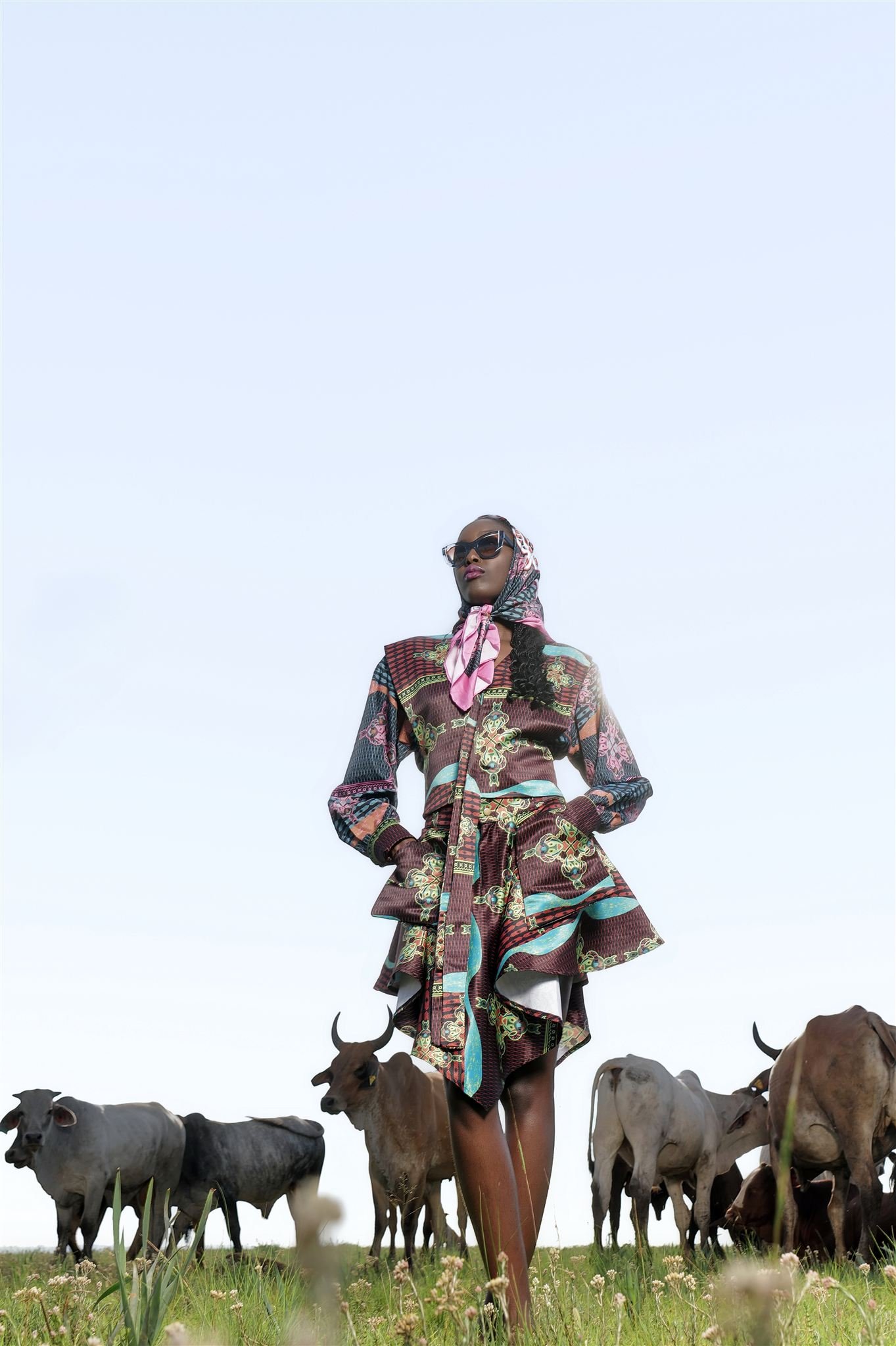 KIM Chakanyuka from AURA Models wearing sunglasses and a colorful, Masamara patterned dress with a headscarf stands in a field of grass with cattle grazing around her.