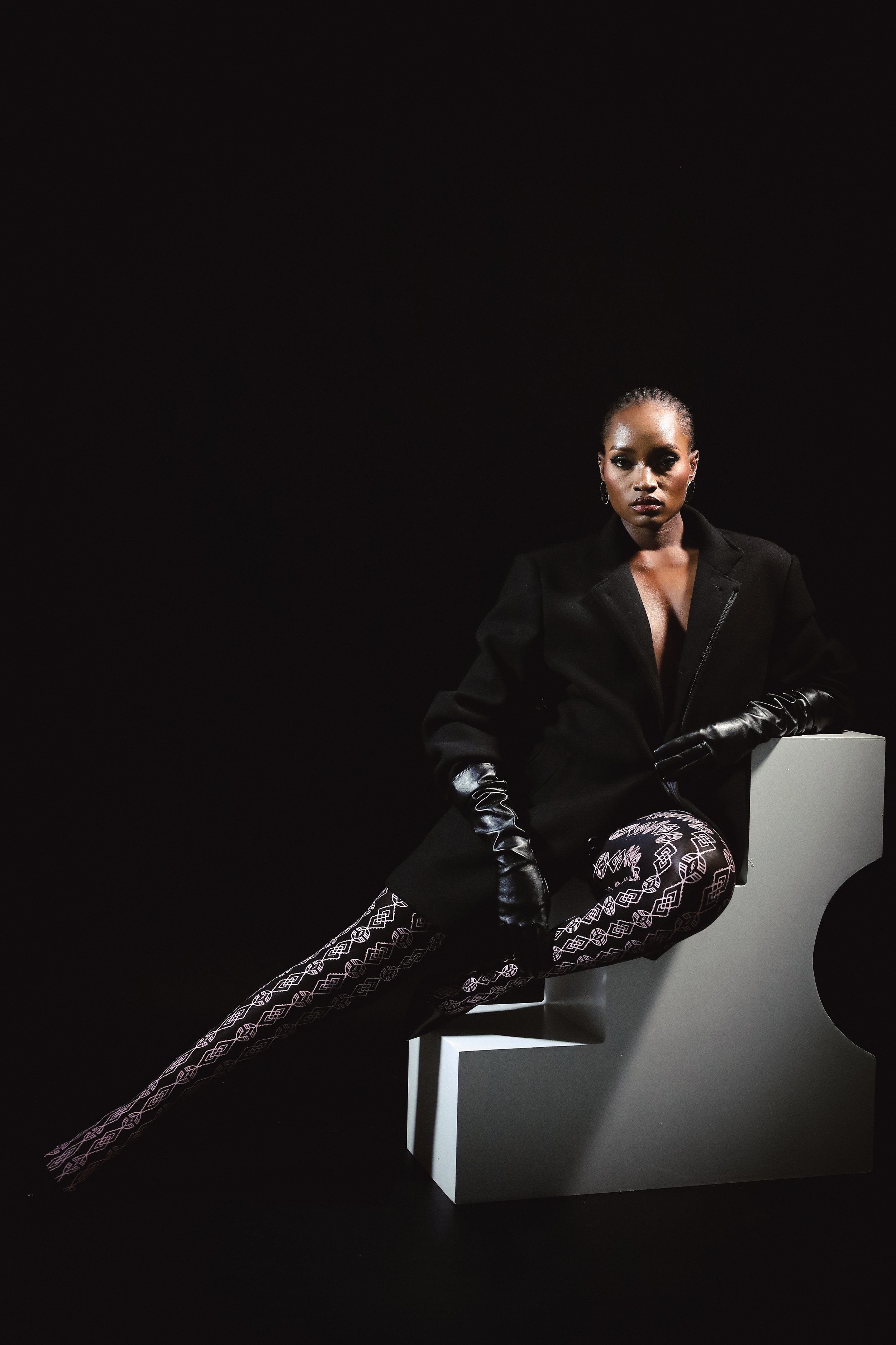 SHALOM Musvosvi from AURA Models with dark skin and slicked-back hair sitting on a white chair against a black background. She is wearing a black blazer, patterned pants, and black leather gloves, with a serious expression.