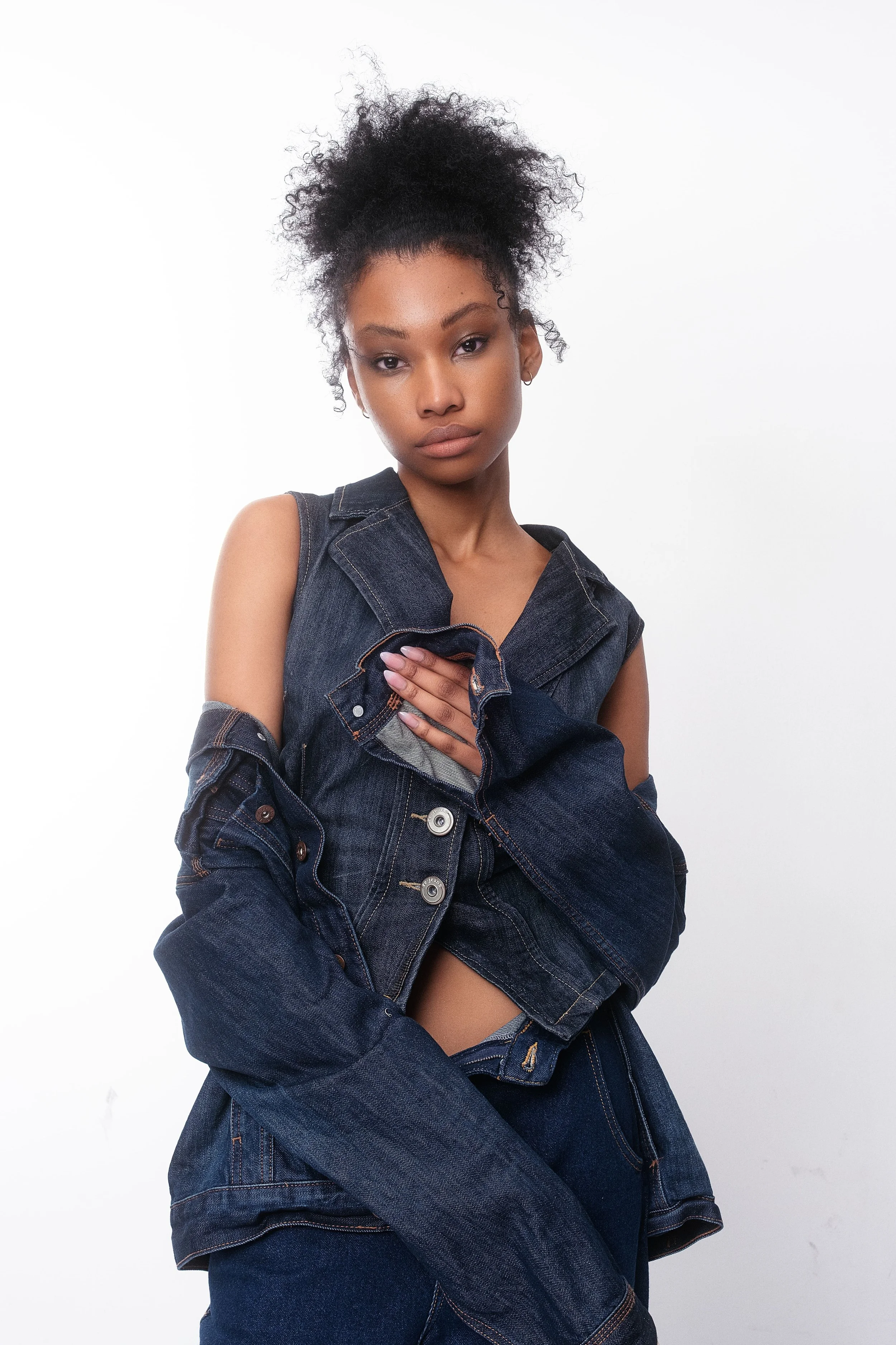 QUISHA Rapoo from AURA Models with curly hair in a high puff hairstyle, wearing a sleeveless denim jacket and jeans, posing against a plain white background.