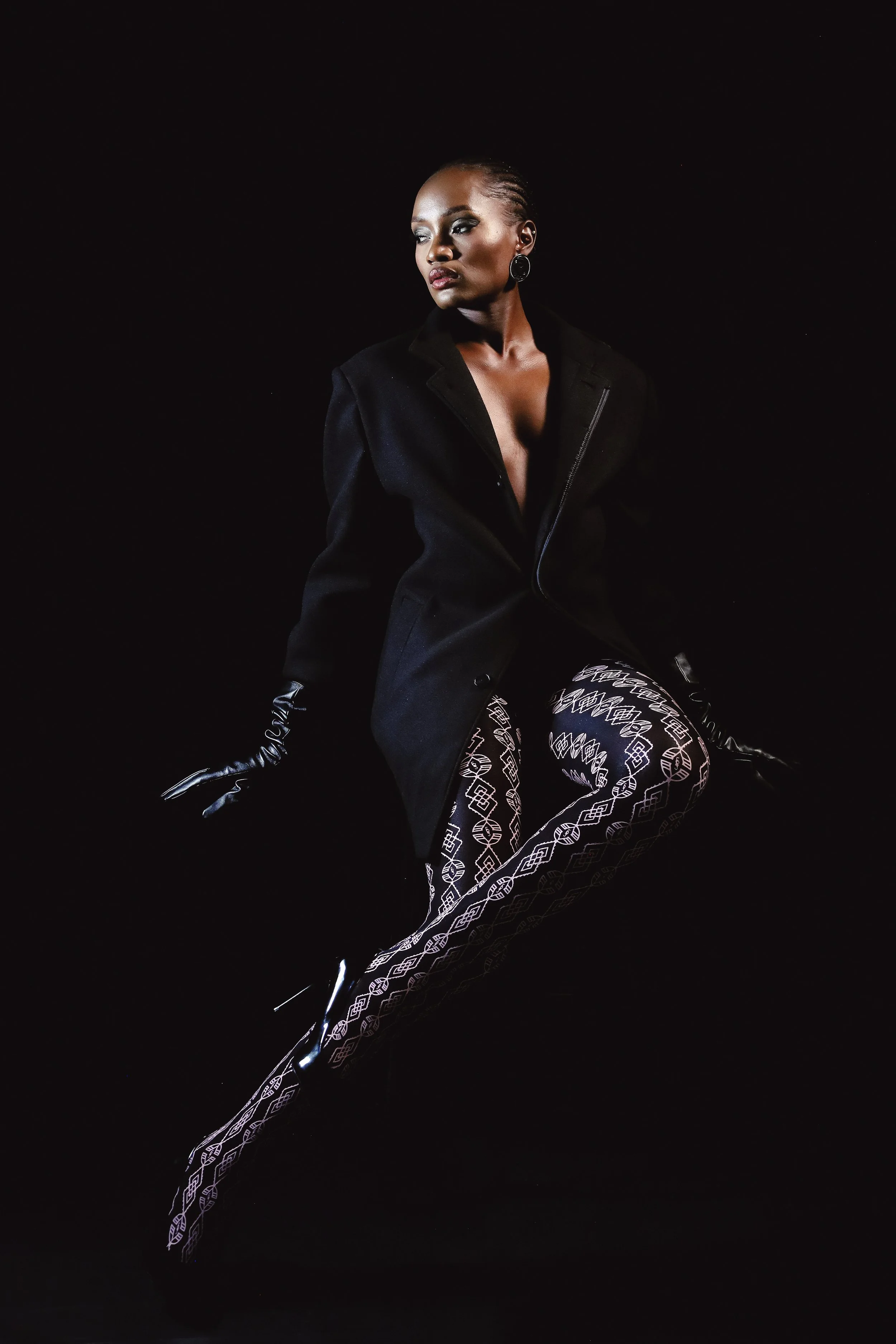 SHALOM Musvosvi from AURA Models with braided hair and dark makeup wearing a black blazer and patterned leggings with a black background.