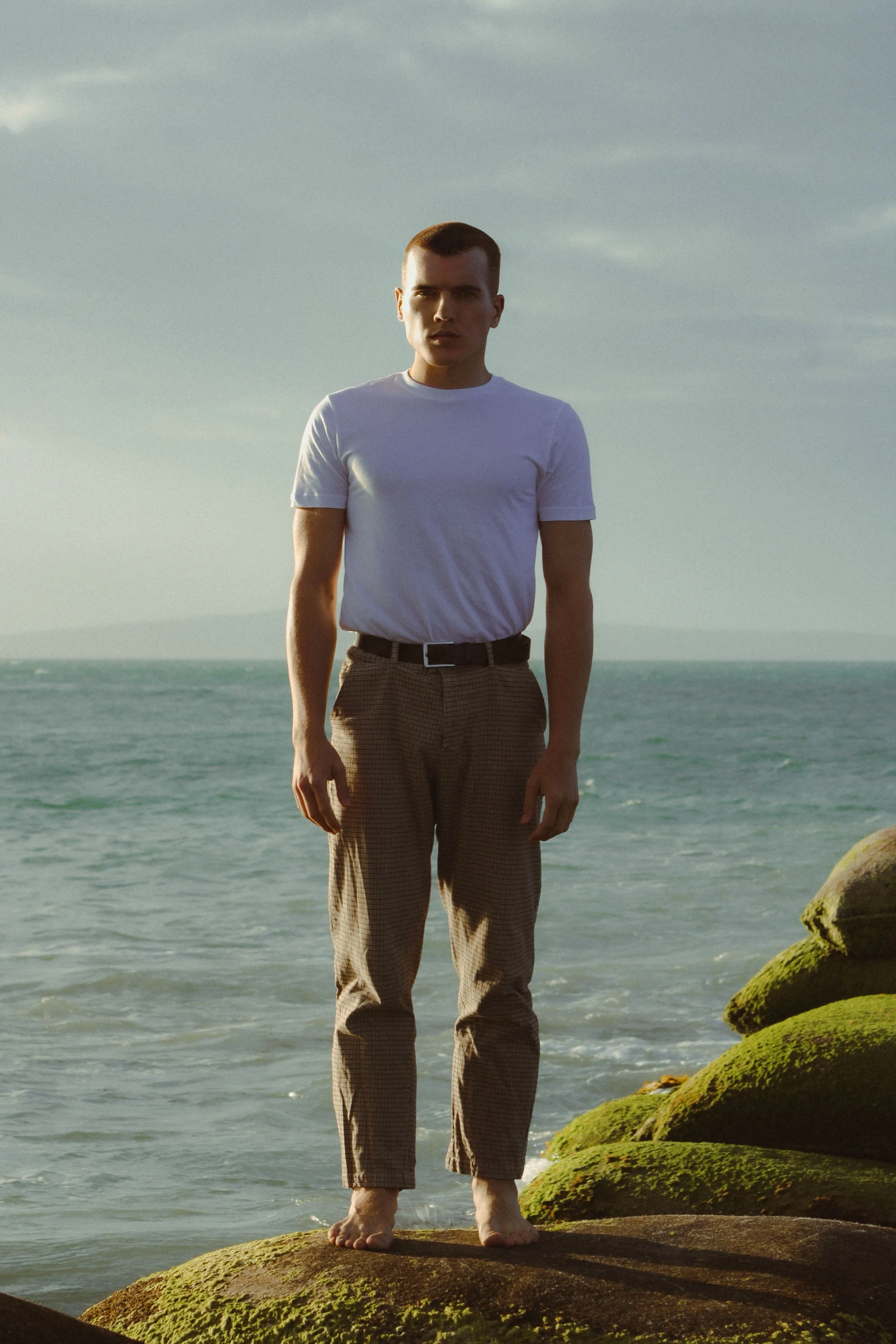 A young man standing on rocks covered in moss by the sea, wearing a white t-shirt and brown checkered pants, with the ocean and sky in the background.
