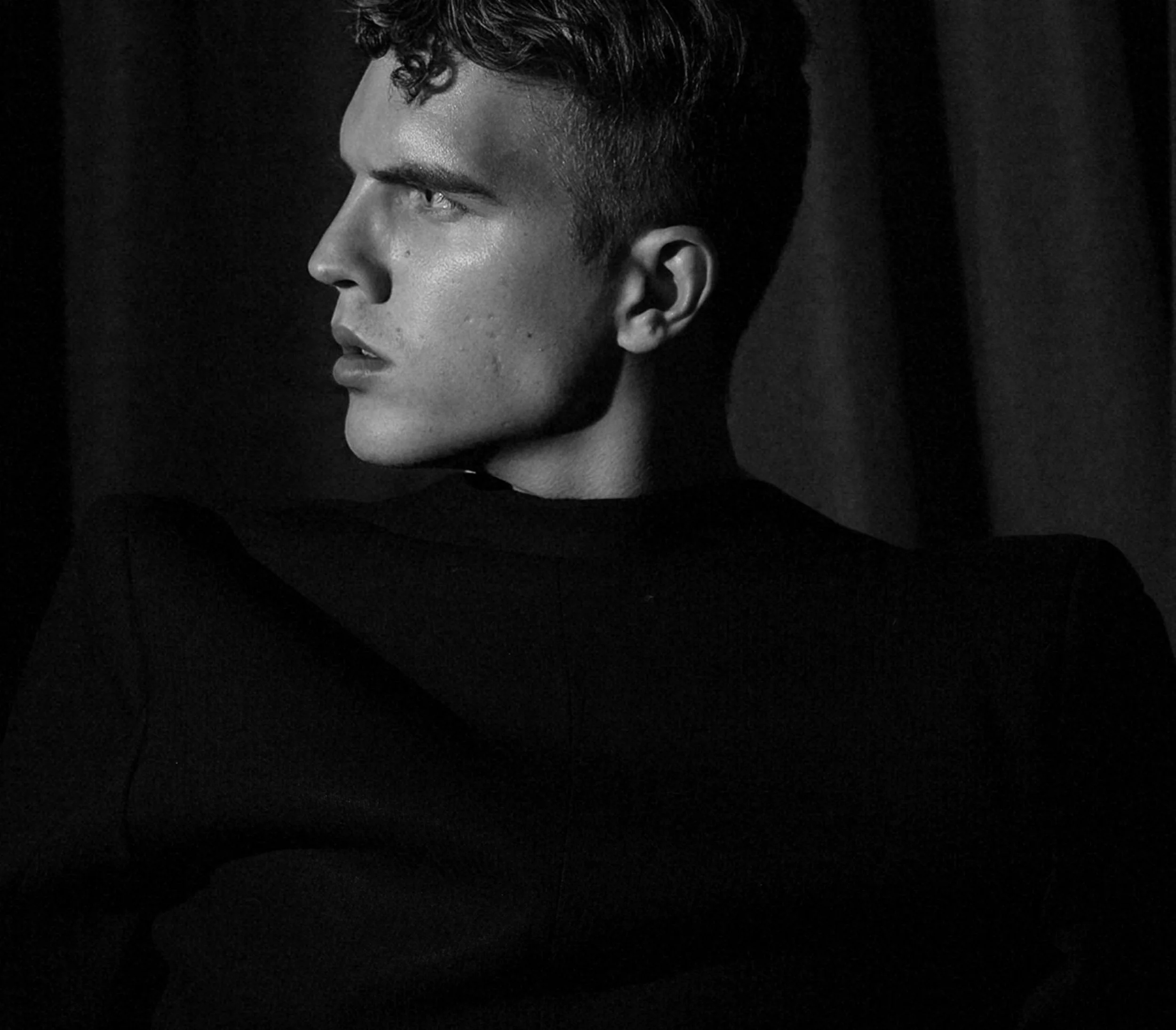 Black and white portrait of a young man with curly hair, gazing intently to the left, against a dark background.