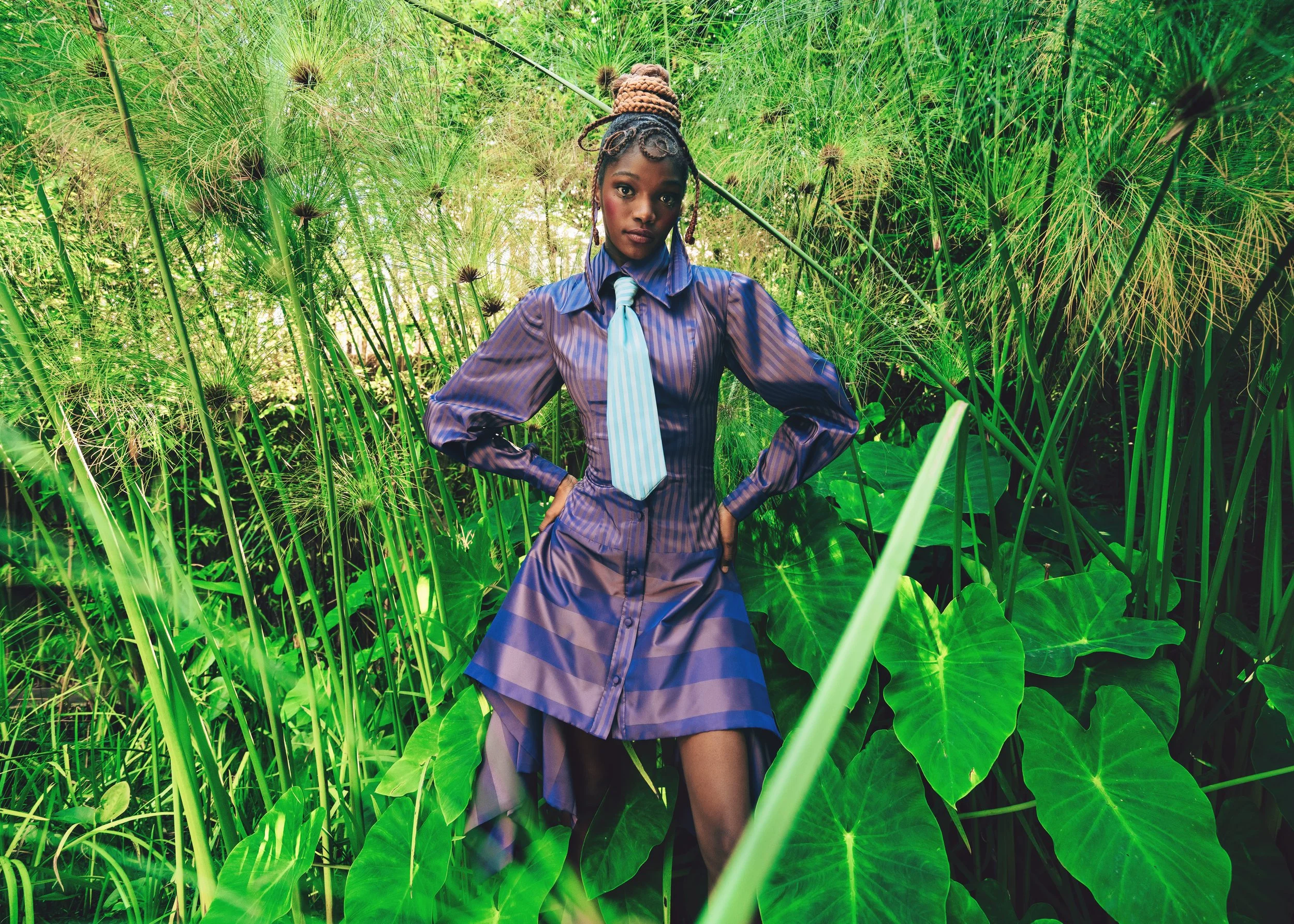 LESEGO Ngidi from AURA Models with dark skin, braided hair styled in an updo, wearing a purple-blue striped shirt, a light blue tie, and a matching skirt, standing amidst tall green plants and papaya leaves in a lush, verdant environment.