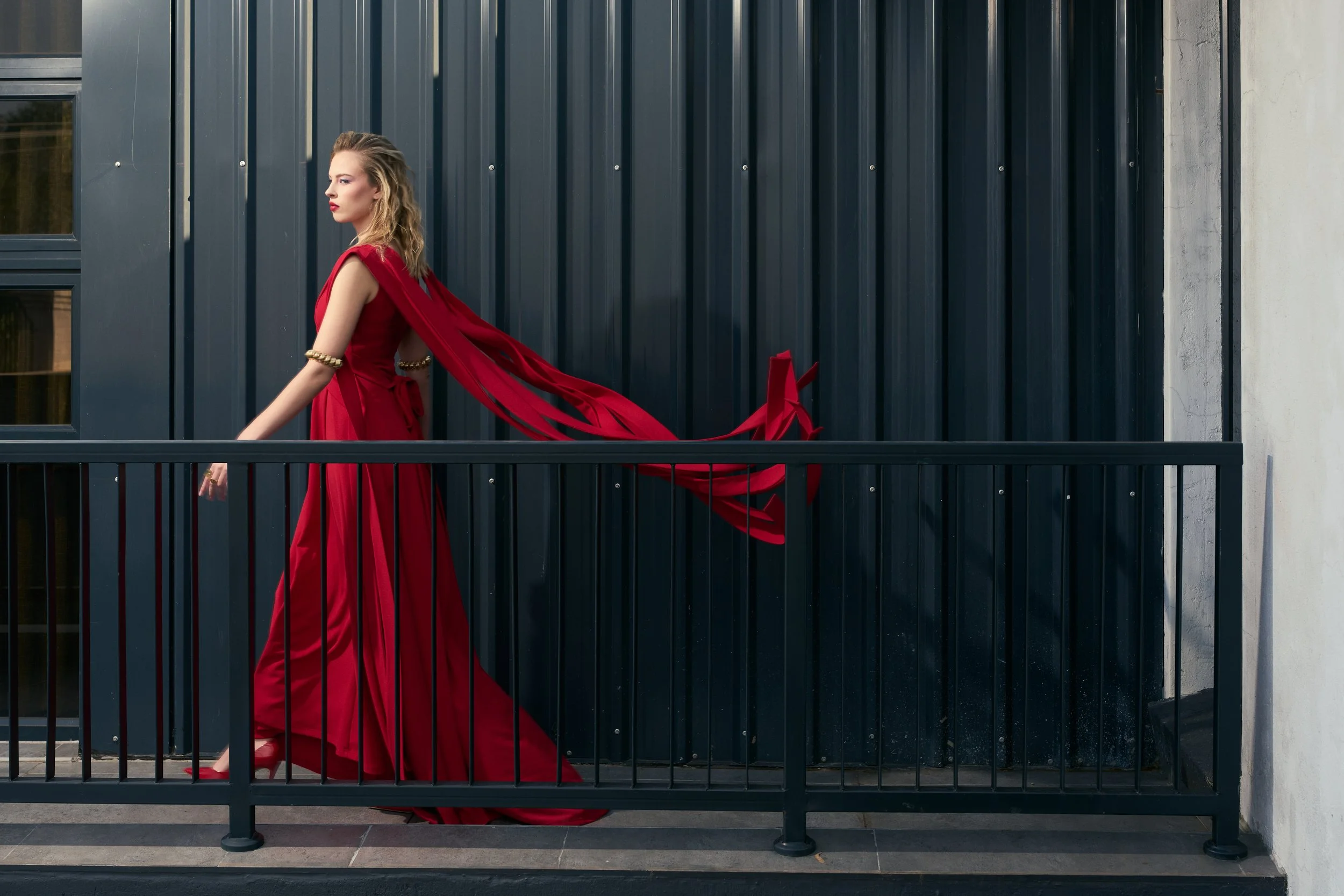 GAIA Fernandez from AURA Models in a long, flowing red dress standing on a balcony with a black metal railing, facing left with a serious expression, against a modern building with dark vertical siding.
