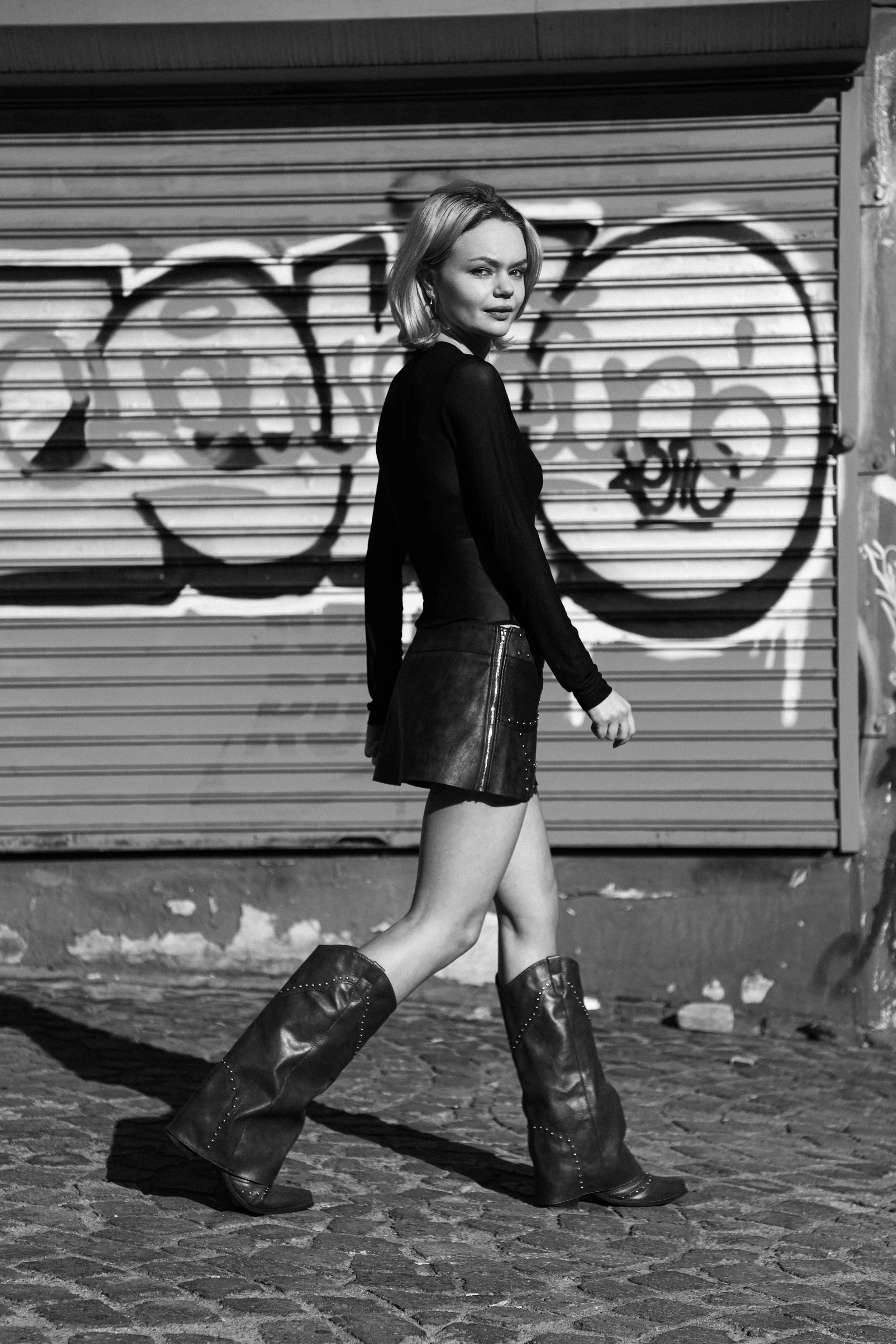 A woman with blonde hair, dressed in a black top and a leather skirt, is walking on cobblestone street in front of a graffiti-covered metal storefront shutter, wearing large boots, in black and white photo.