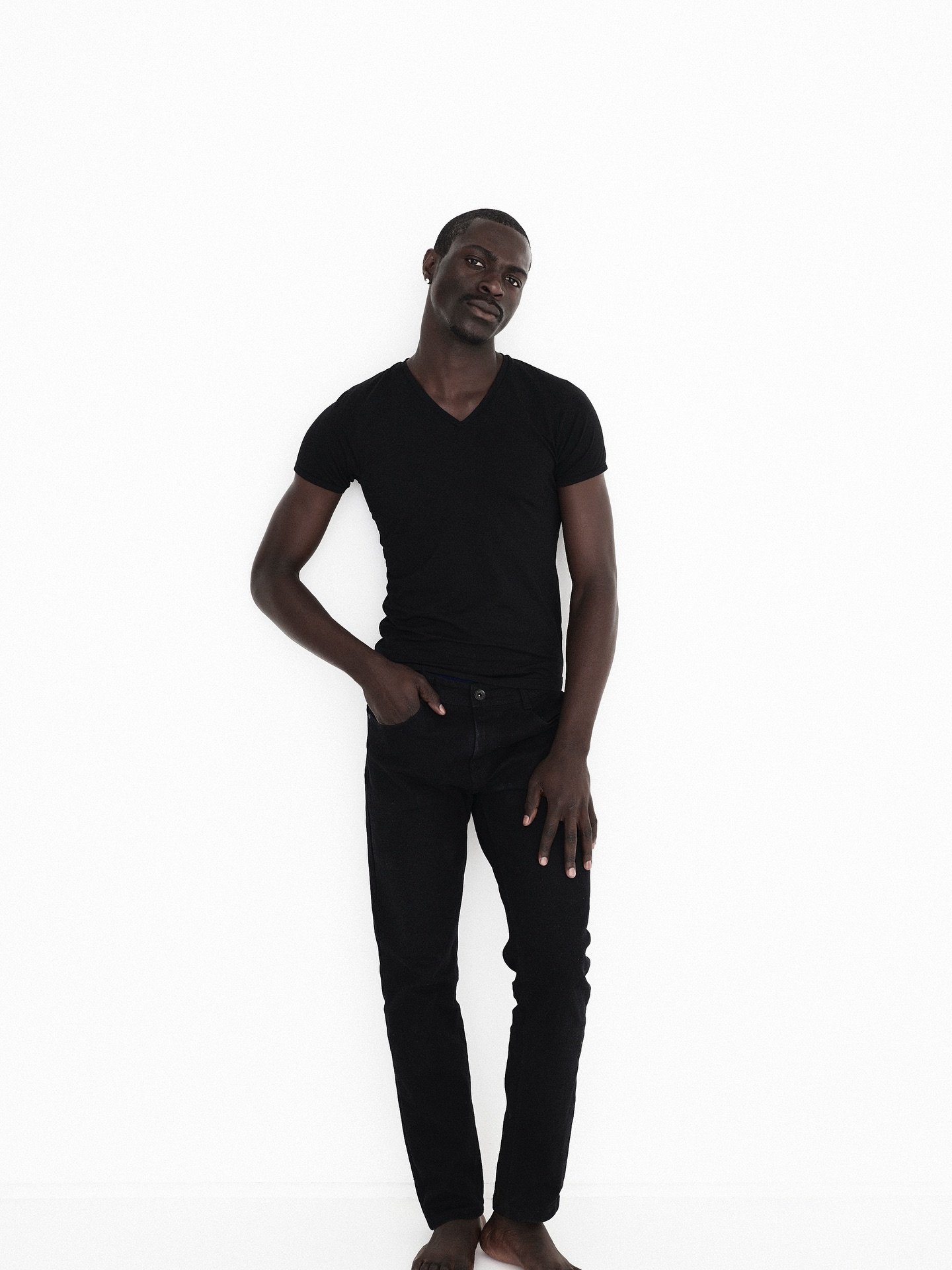 A young man with dark skin standing barefoot against a plain white background, wearing a black V-neck T-shirt and black jeans, with one hand in his pocket and the other resting on his thigh.