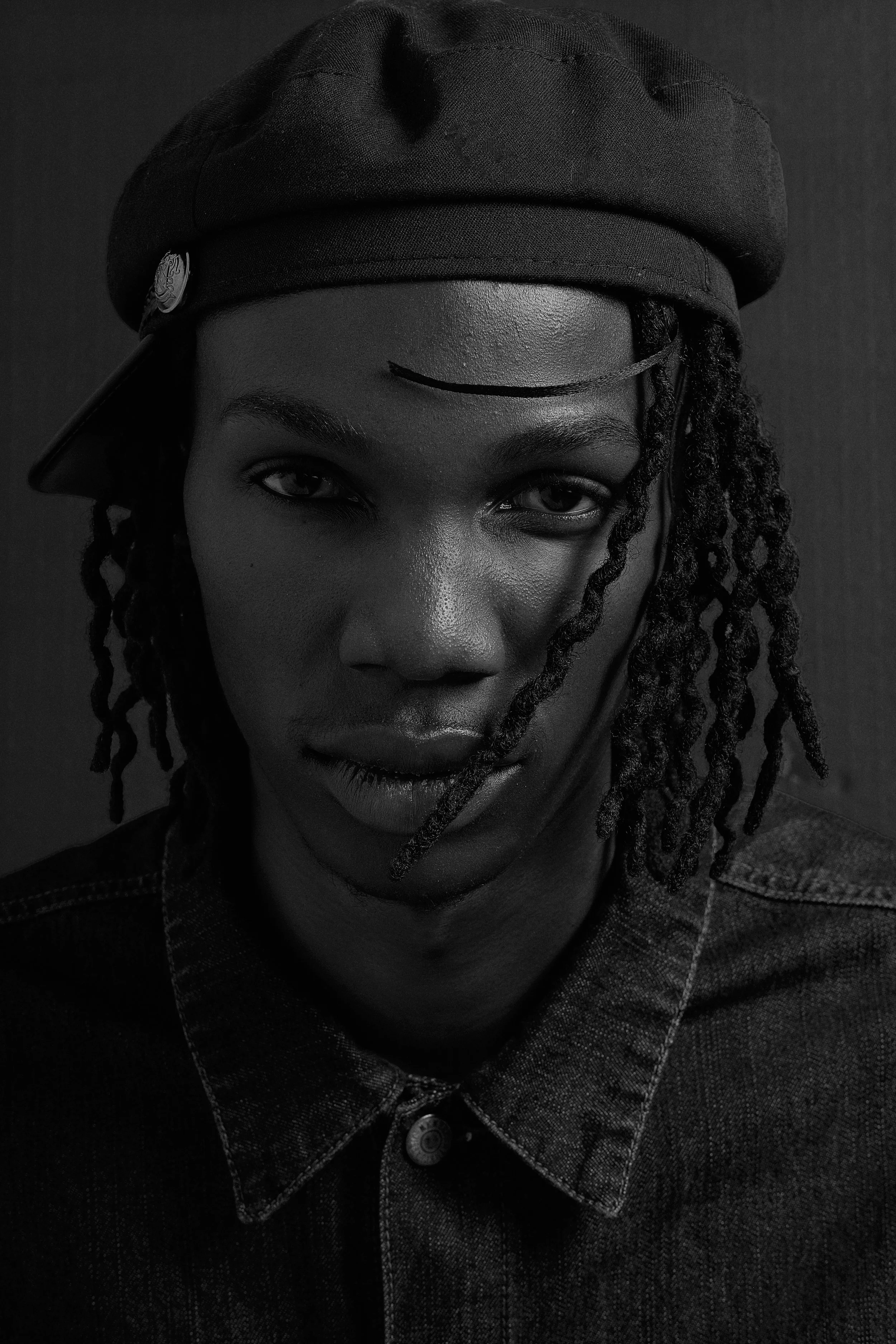 Black and white portrait of TSHEDZA Ramavheya from AURA Models wearing a dark beret tilted to the side, with dreadlocks and a button-up denim jacket.