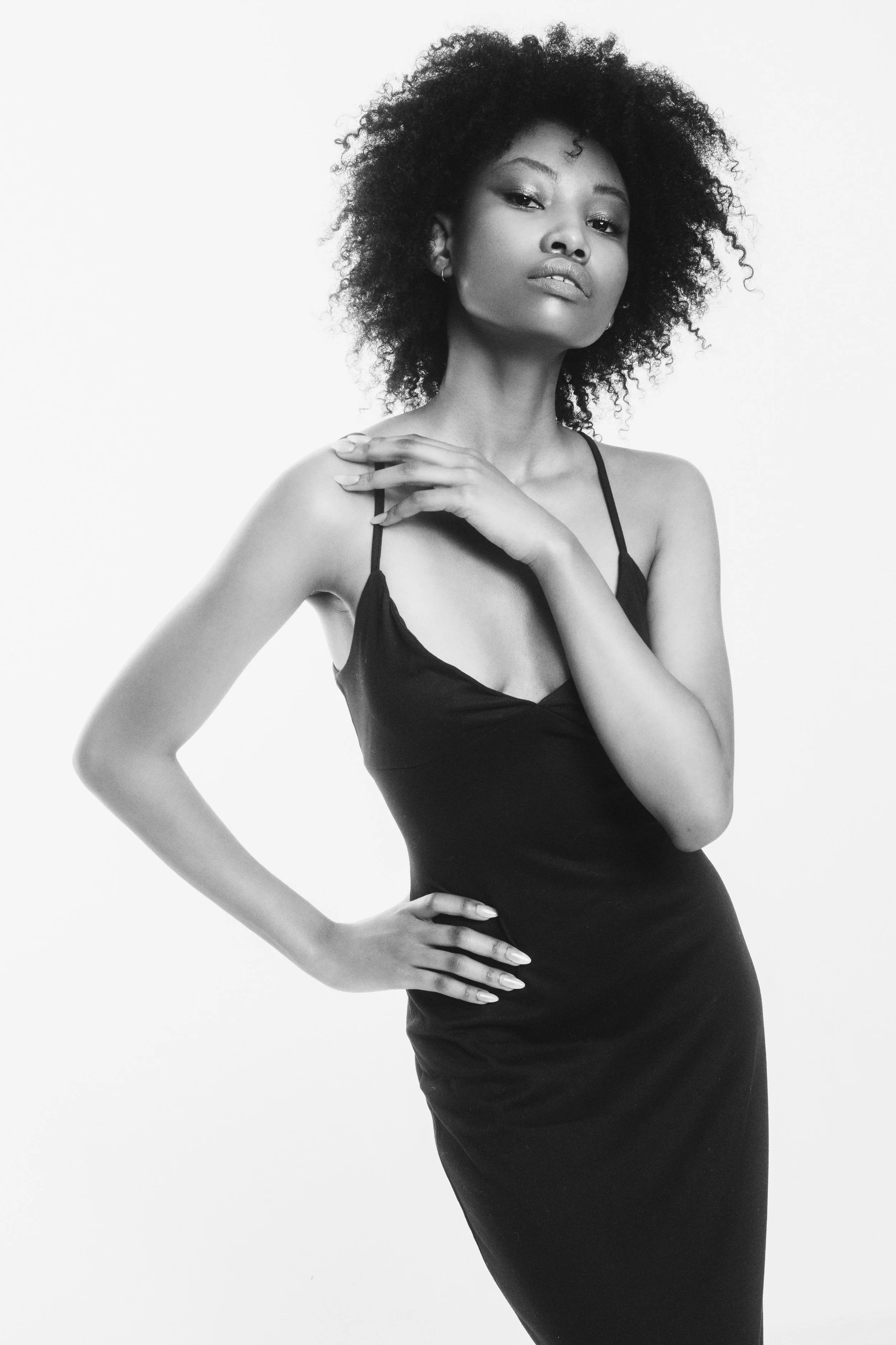 Black and white photo of QUISHA Rapoo from AURA Models with curly hair wearing a spaghetti strap dress, posing with one hand on her hip and the other resting on her shoulder, against a plain light background.