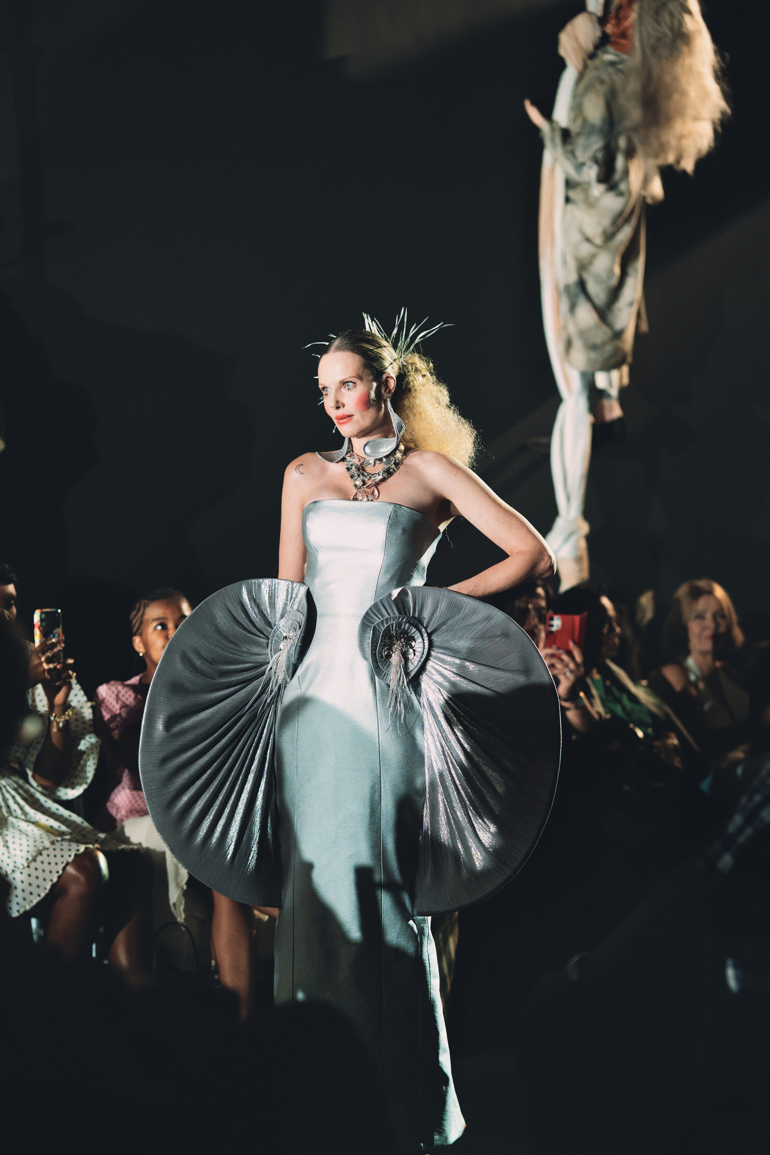 NATALIA Kostanova from AURA Models walking on a runway in a silver, metallic gown with large, circular, pleated details. She has bright makeup, an elaborate hairstyle, and wears statement jewelry, with an audience watching.