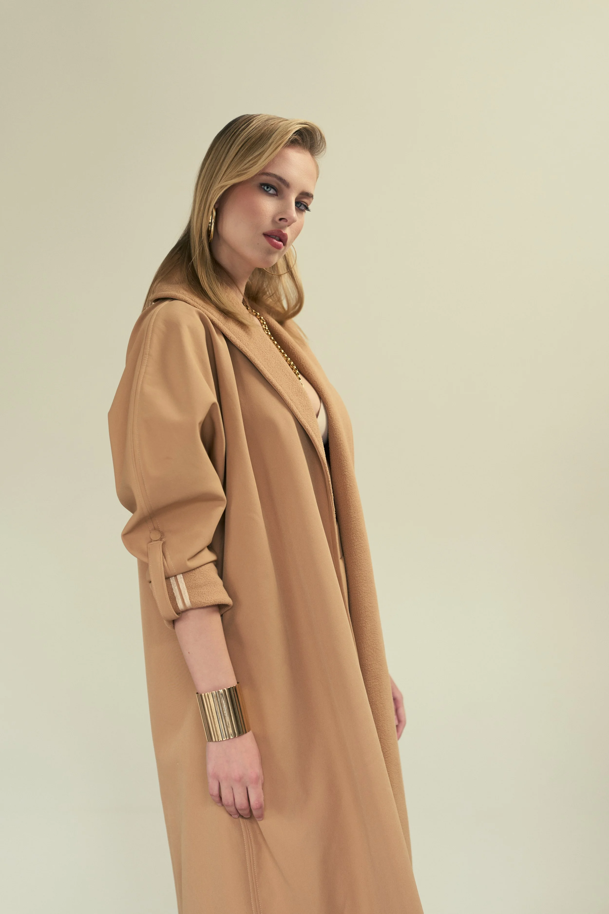GAIA Fernandez from AURA Models with blonde hair, wearing a beige coat, gold hoop earrings, and a gold bracelet, standing against a plain light yellow background.