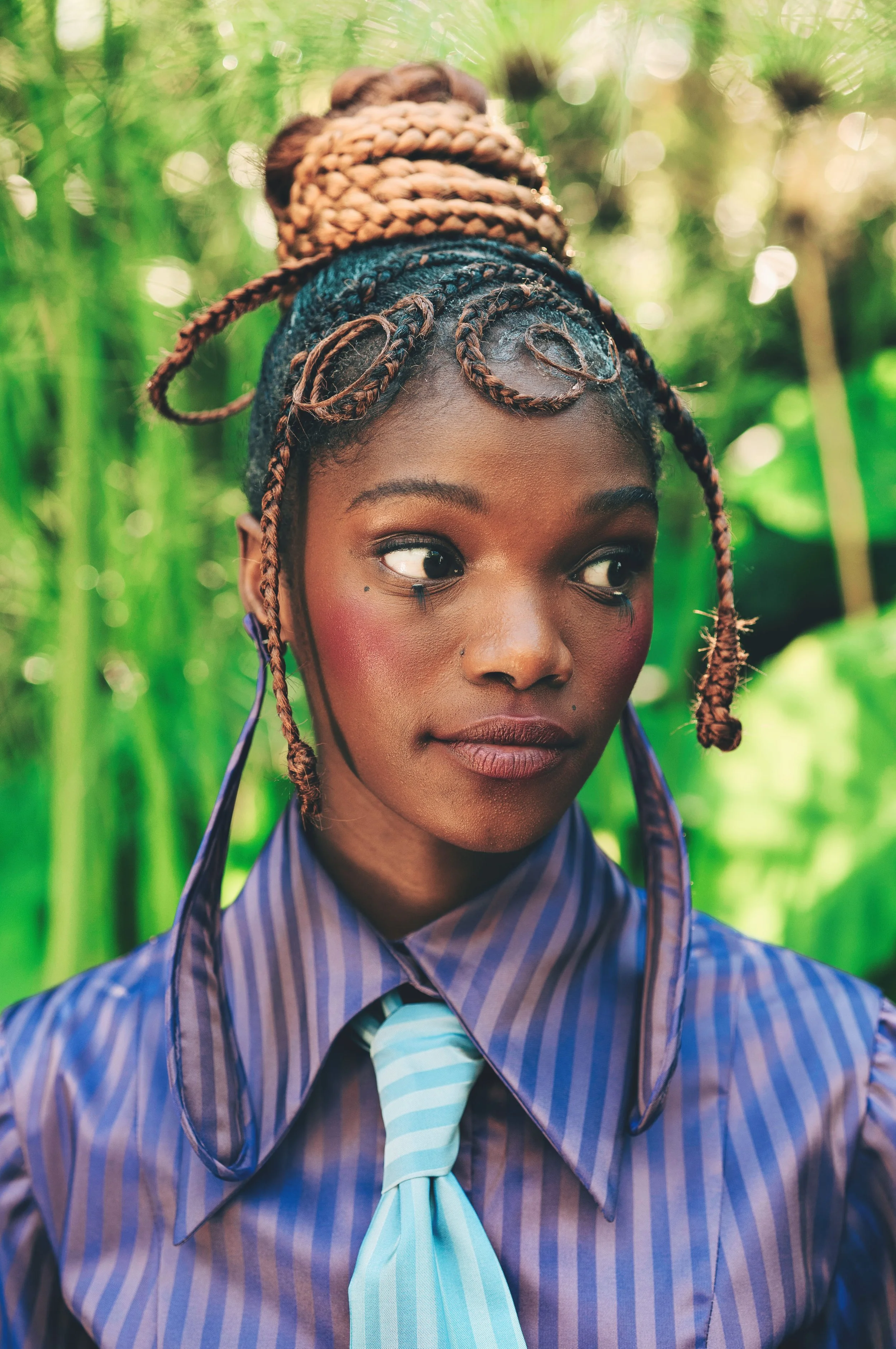 Close-up portrait of LESEGO Ngidi from AURA Models with braided hair styled in an updo, wearing a blue striped shirt and matching tie, with green foliage background.
