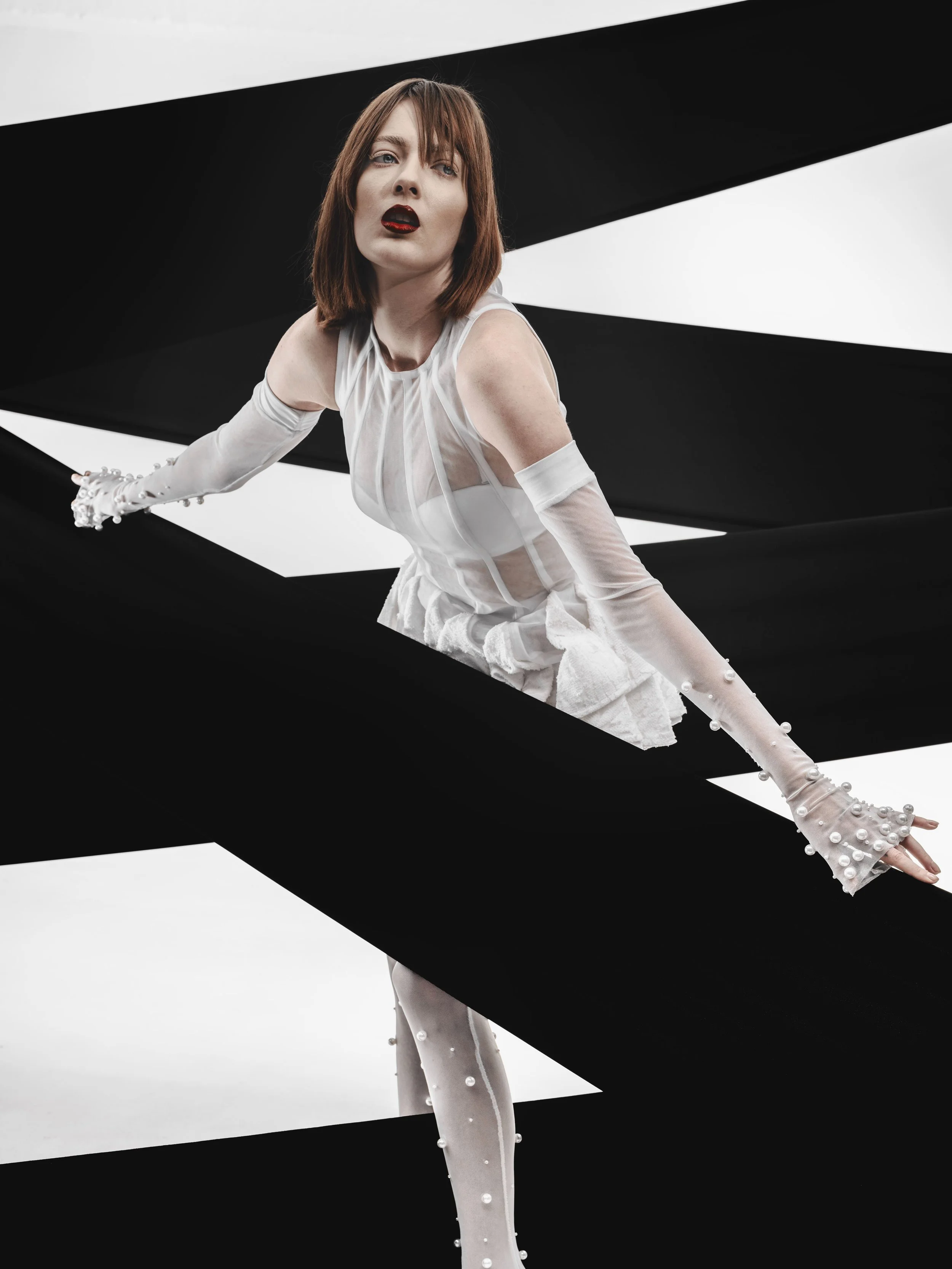 CAYLEIGH Bancroft from AURA Models with short brown hair wearing a white dress and sheer gloves with pearl embellishments, striking a dynamic pose against a minimalist black and white geometric background.