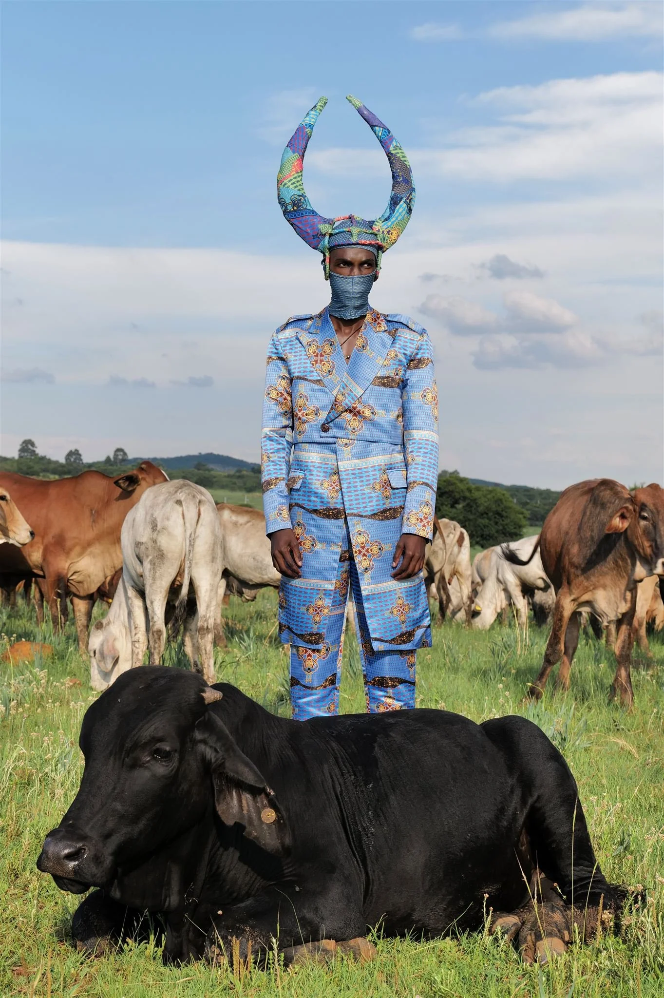 TSHEDZA Ramavheya from AURA Models dressed in colorful Masamara traditional attire with a matching headdress and face mask is standing in a green field with cows. 