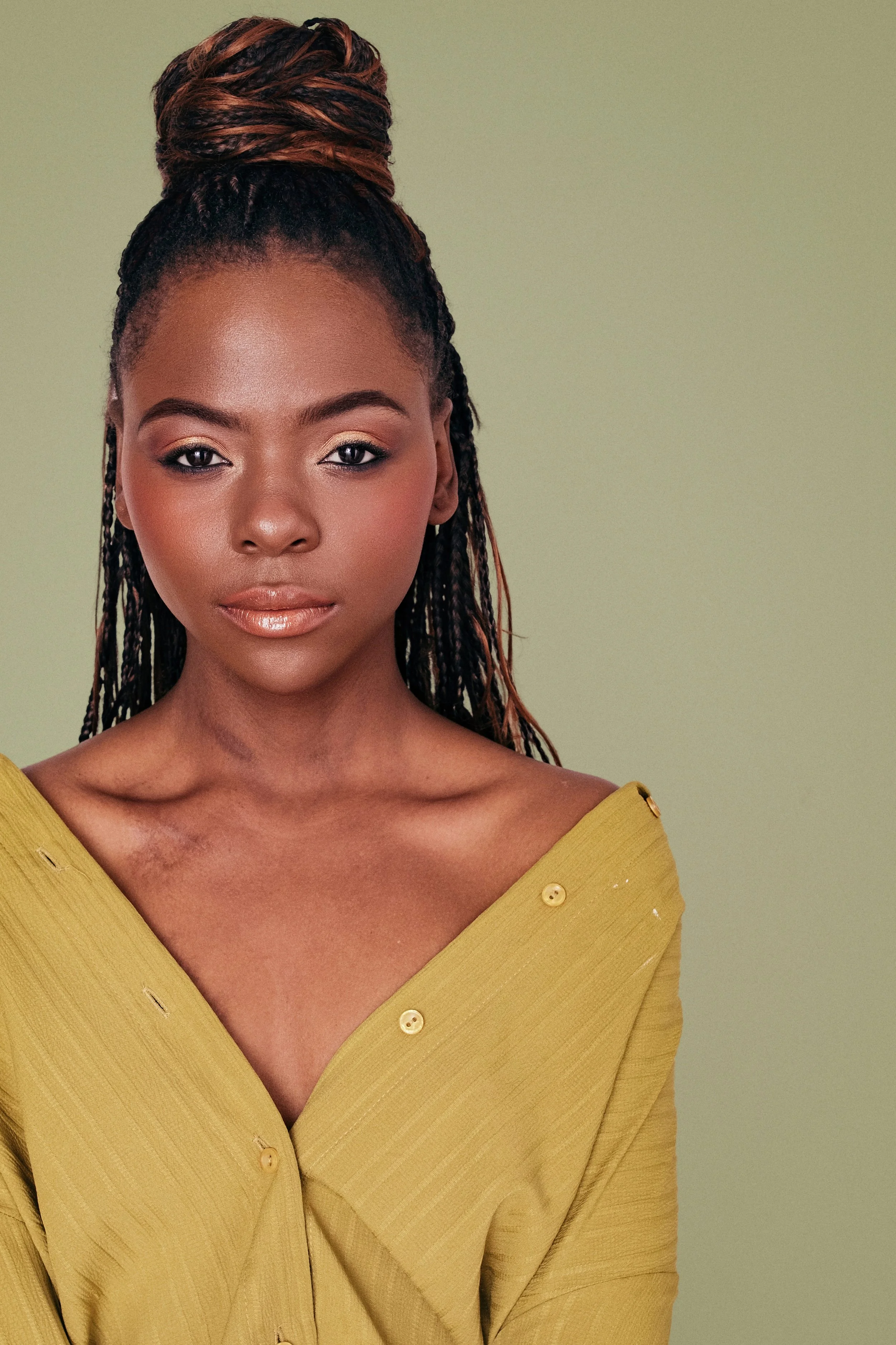 Portrait of a woman with braided hair piled on top, wearing a yellow off-shoulder blouse against a green background.